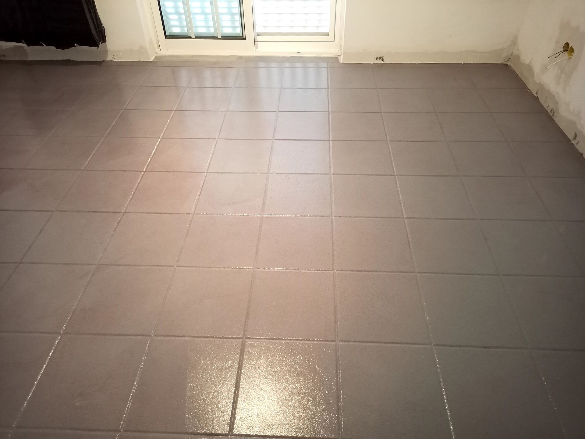 Piso de azulejos cinza com aparência brilhante e úmida, próximo a uma janela e parede.