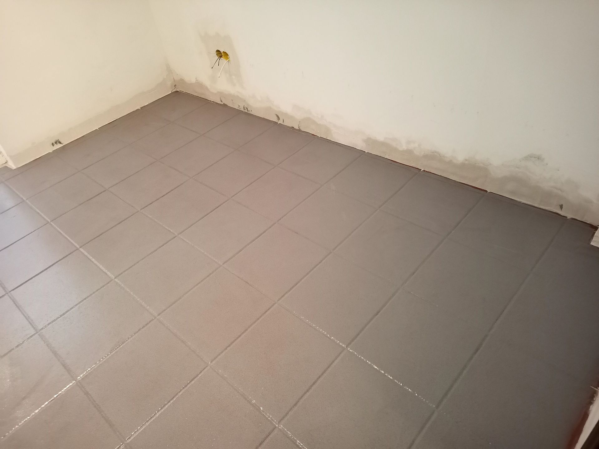 Piso de azulejo cinza em um canto do cômodo, paredes brancas, com tomada elétrica e rodapé.