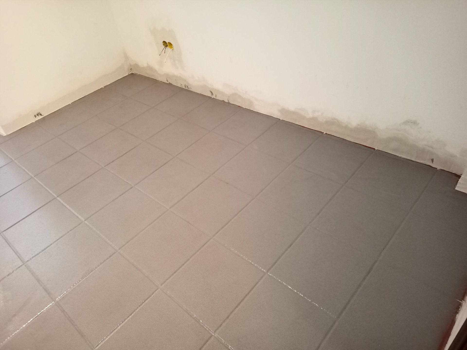 Piso de azulejos cinza em um cômodo vazio, com paredes brancas e uma tomada elétrica visível.