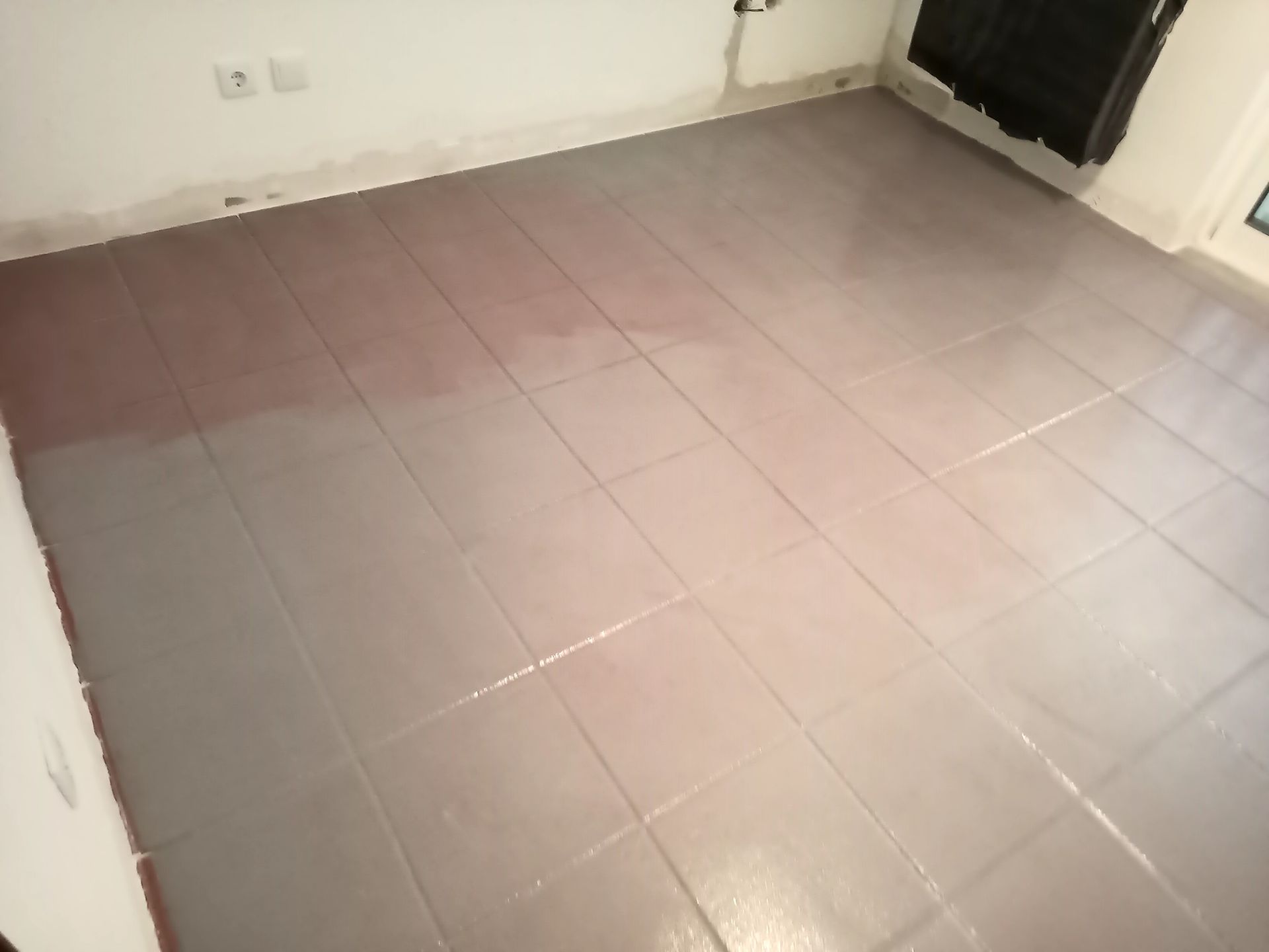 Piso de azulejos cinza com uma área úmida e avermelhada. O cômodo tem paredes brancas e um objeto escuro no canto.