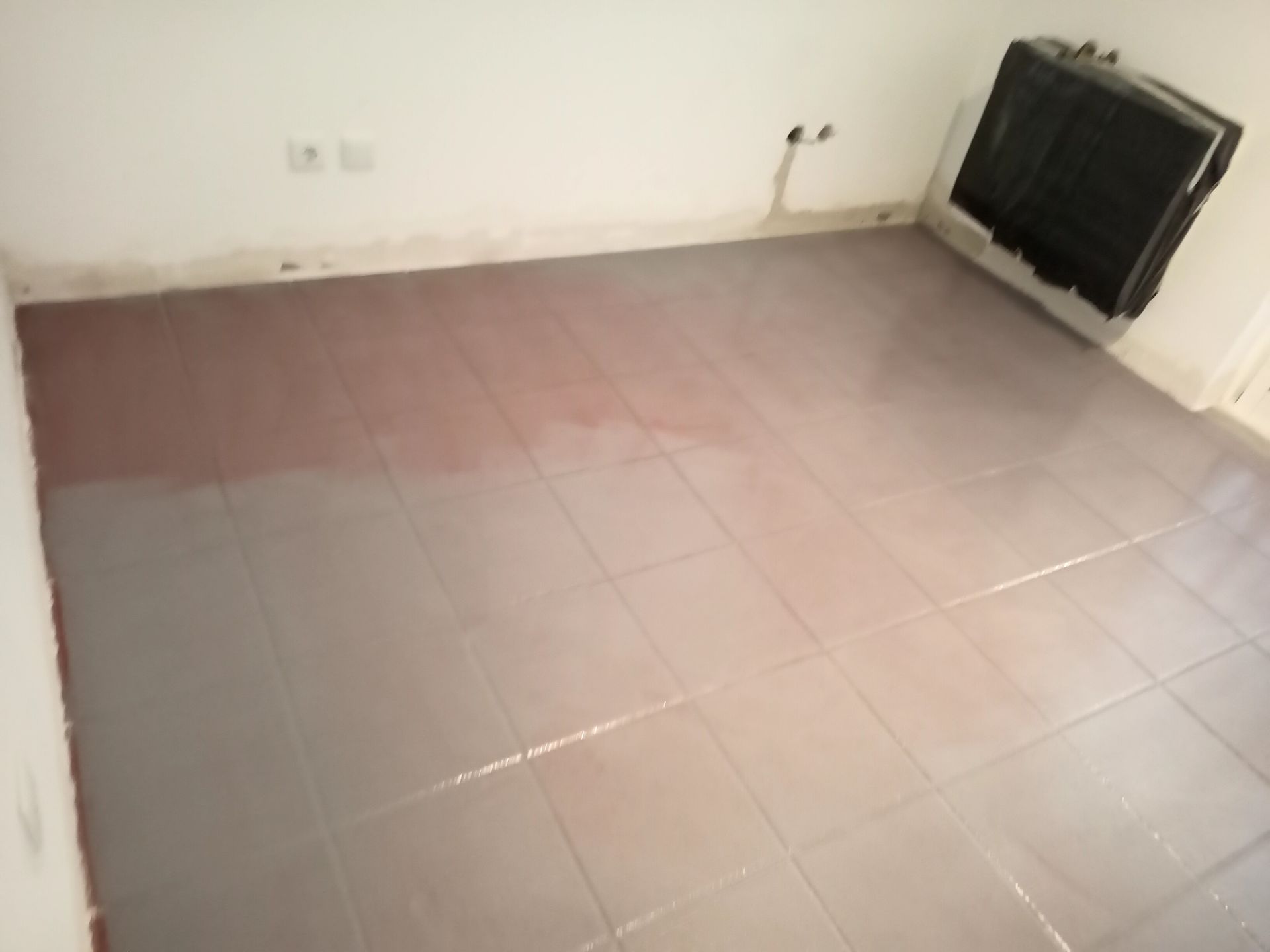 Piso de azulejo cinza em um cômodo, com uma seção parcialmente pintada de marrom-avermelhado perto da parede. Um aquecedor está no canto.