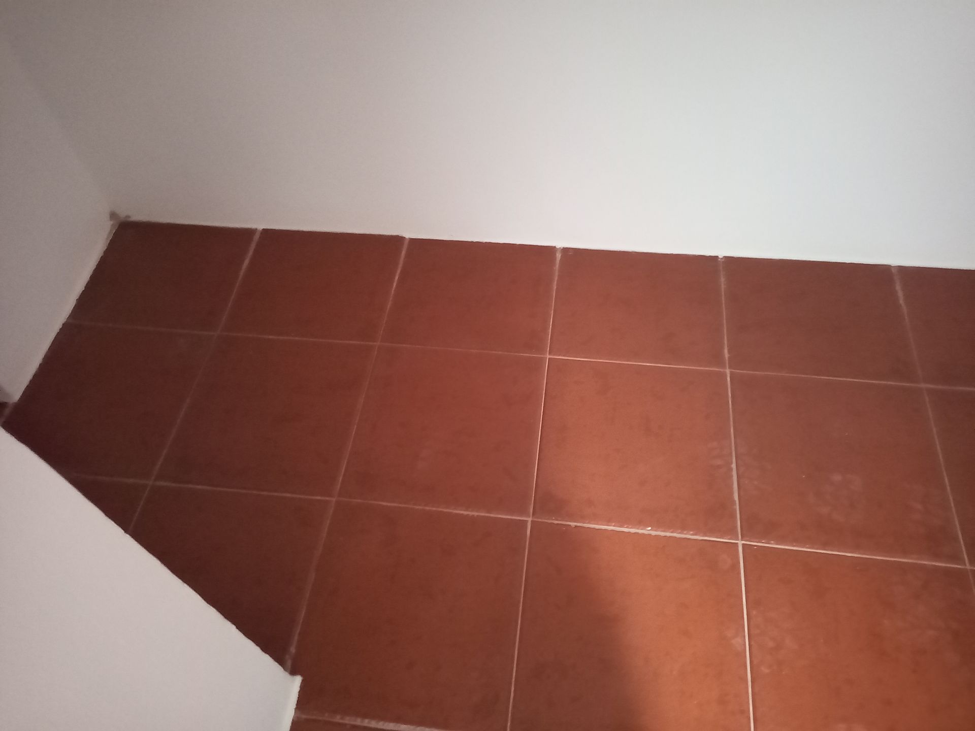 Piso de ladrilho marrom no canto de uma sala, adjacente a paredes brancas.
