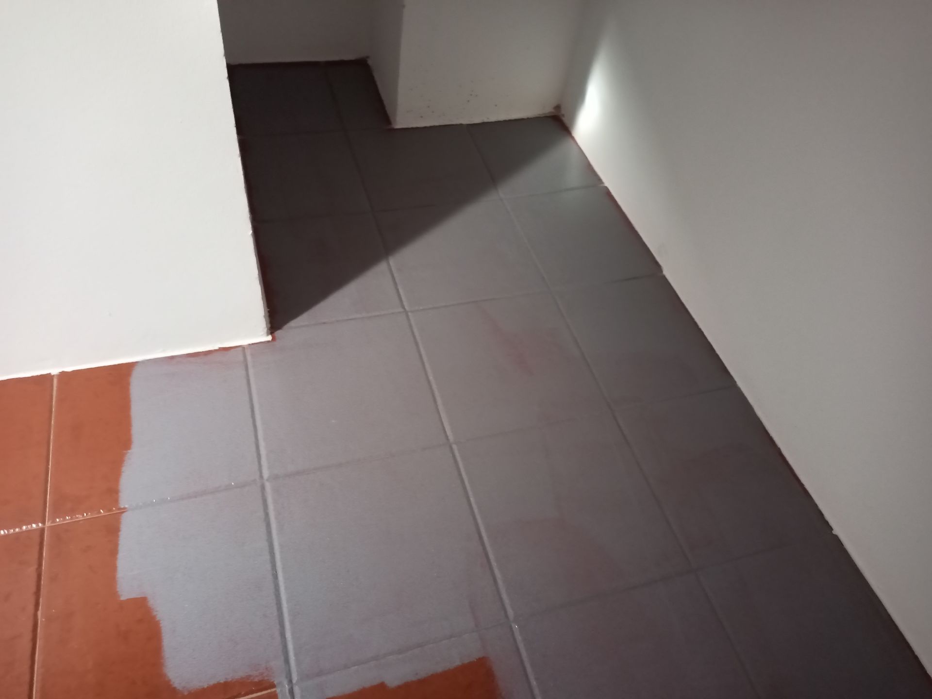 Piso de ladrilhos vermelhos e cinza com paredes brancas; luz natural projetando sombras.