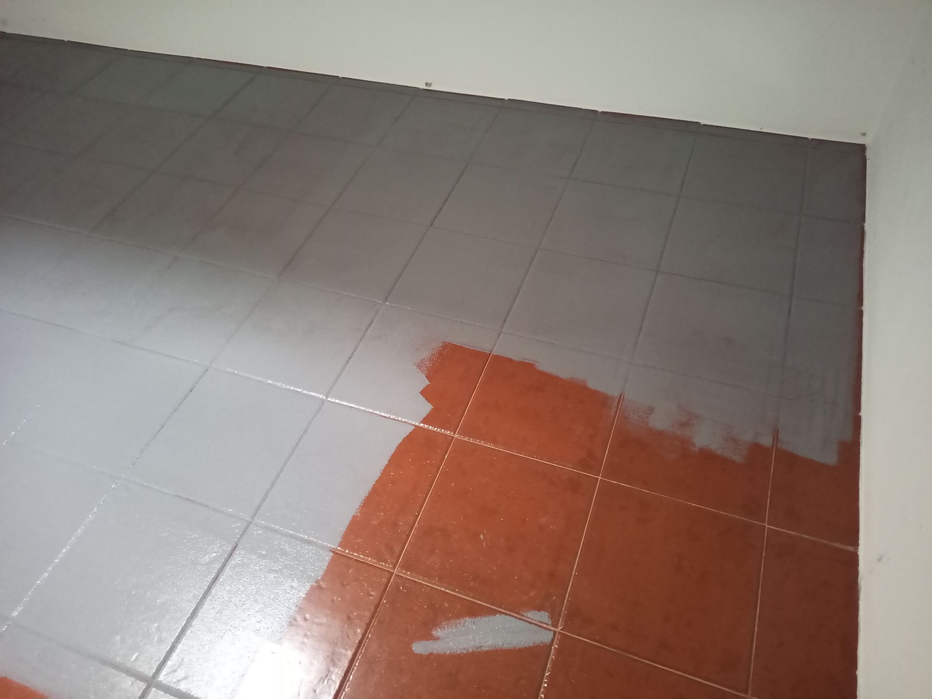 Piso de ladrilhos pintados metade de cinza e metade de laranja sem pintura, ao lado de uma parede branca.