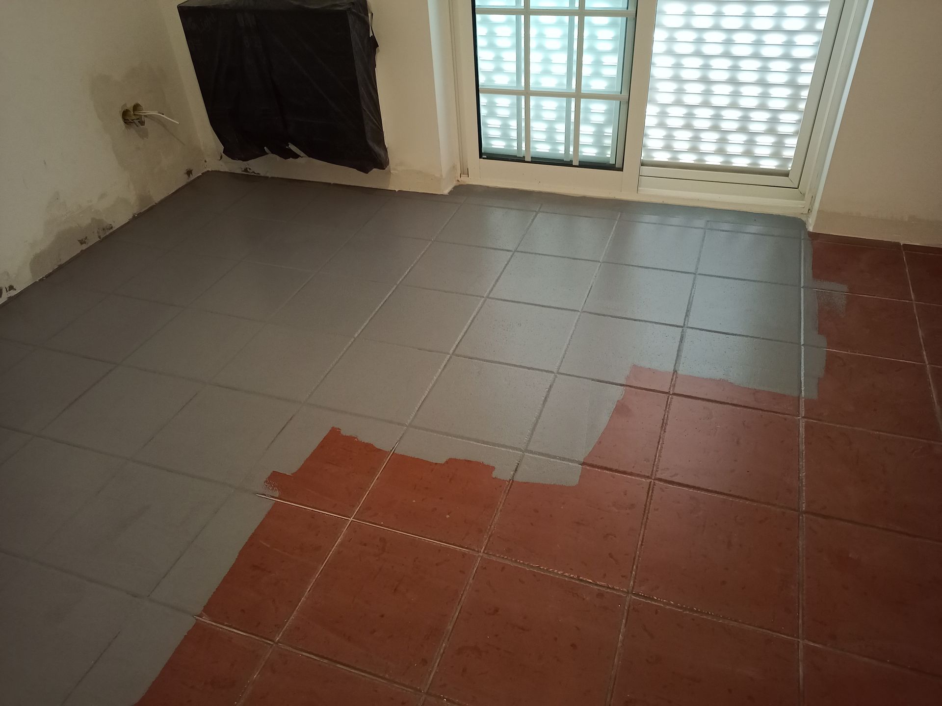 Quarto com piso de ladrilhos pintados de cinza e ladrilhos marrom-avermelhados sem pintura, próximo a uma janela e parede.