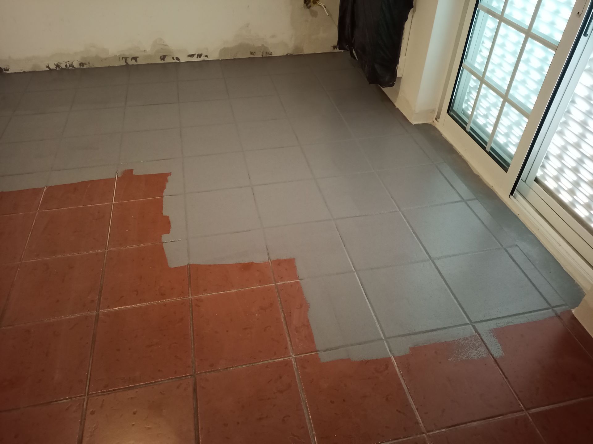 Azulejos no chão, parcialmente pintados de cinza, ao lado de uma janela com venezianas brancas.