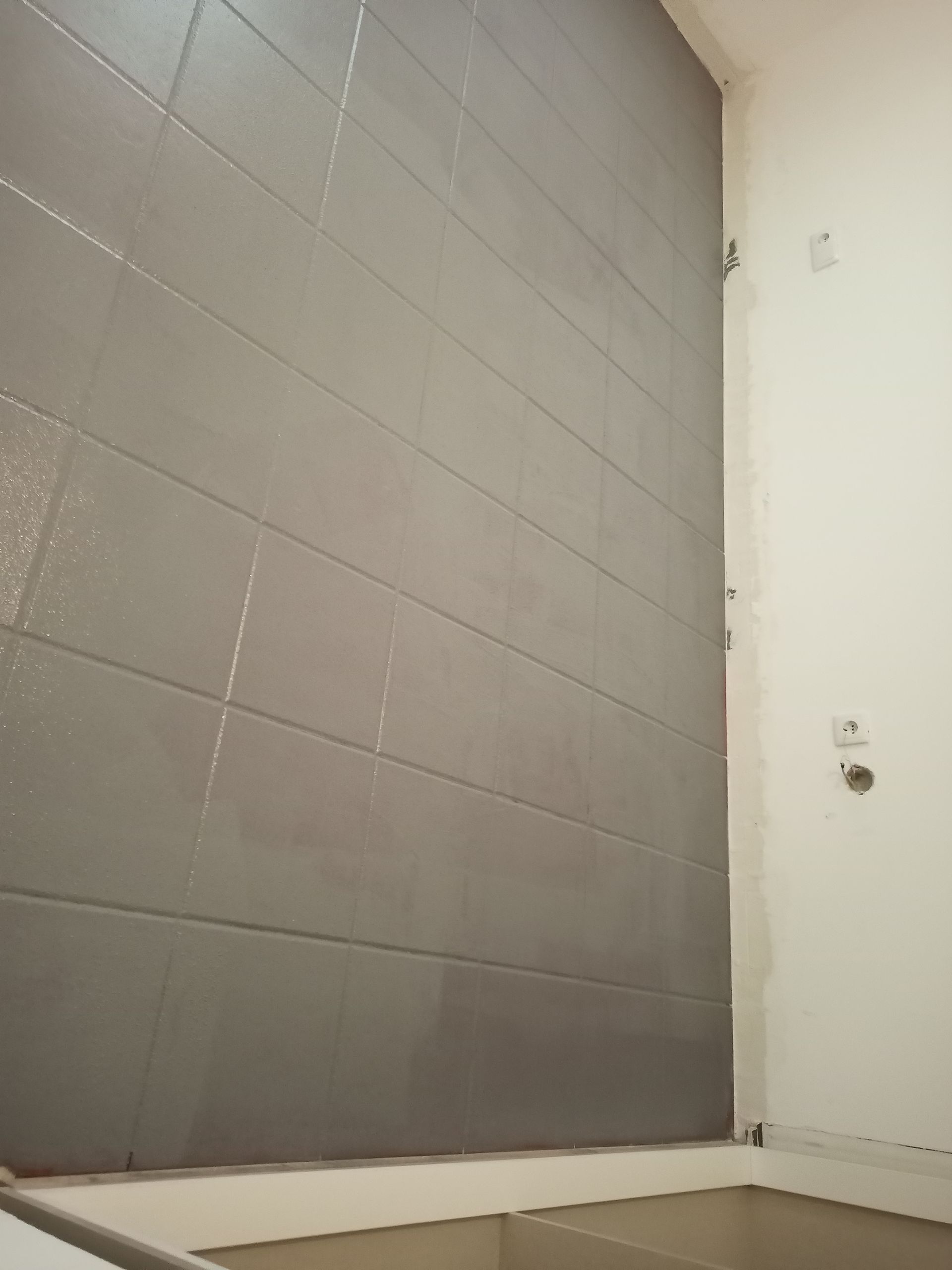 Parede de azulejos pintada de cinza em um banheiro, com uma parede branca à direita e vista parcial de uma prateleira.