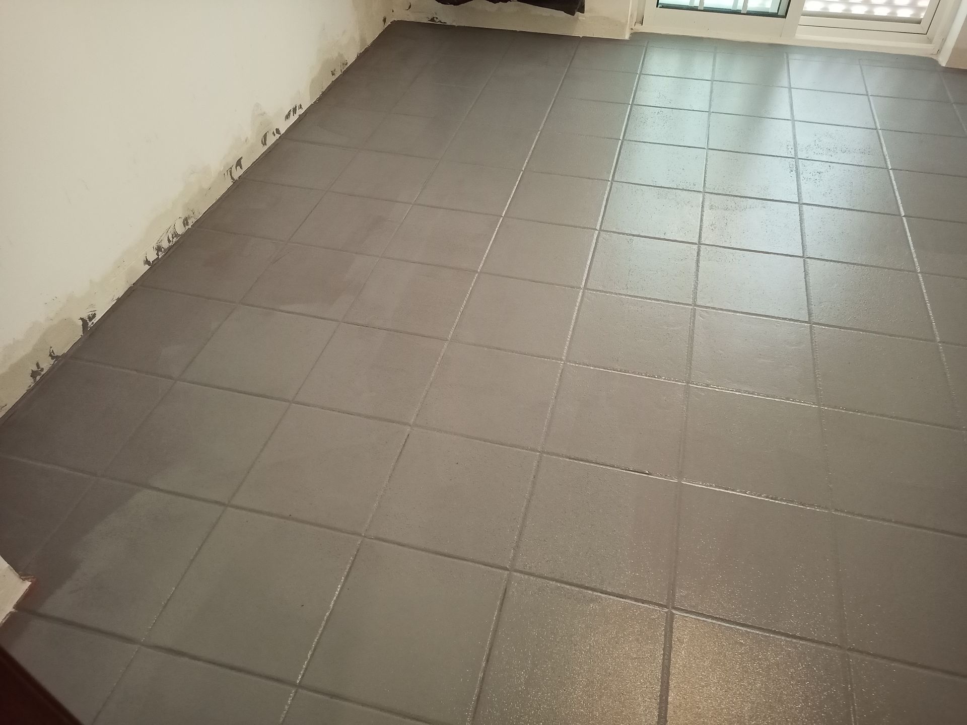Piso de ladrilhos cinza parcialmente coberto por uma camada de cinza mais escuro, próximo a uma parede branca e uma janela.