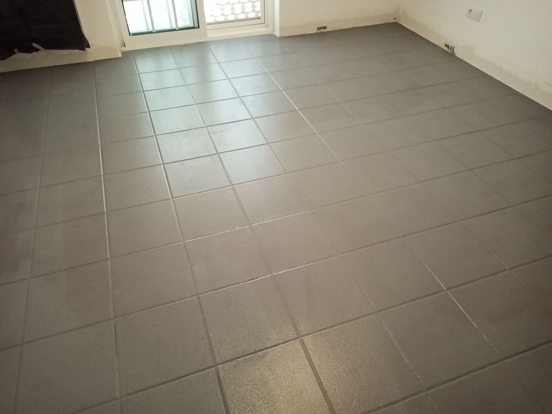 Piso de azulejos cinza em um cômodo vazio, com janela e paredes brancas.