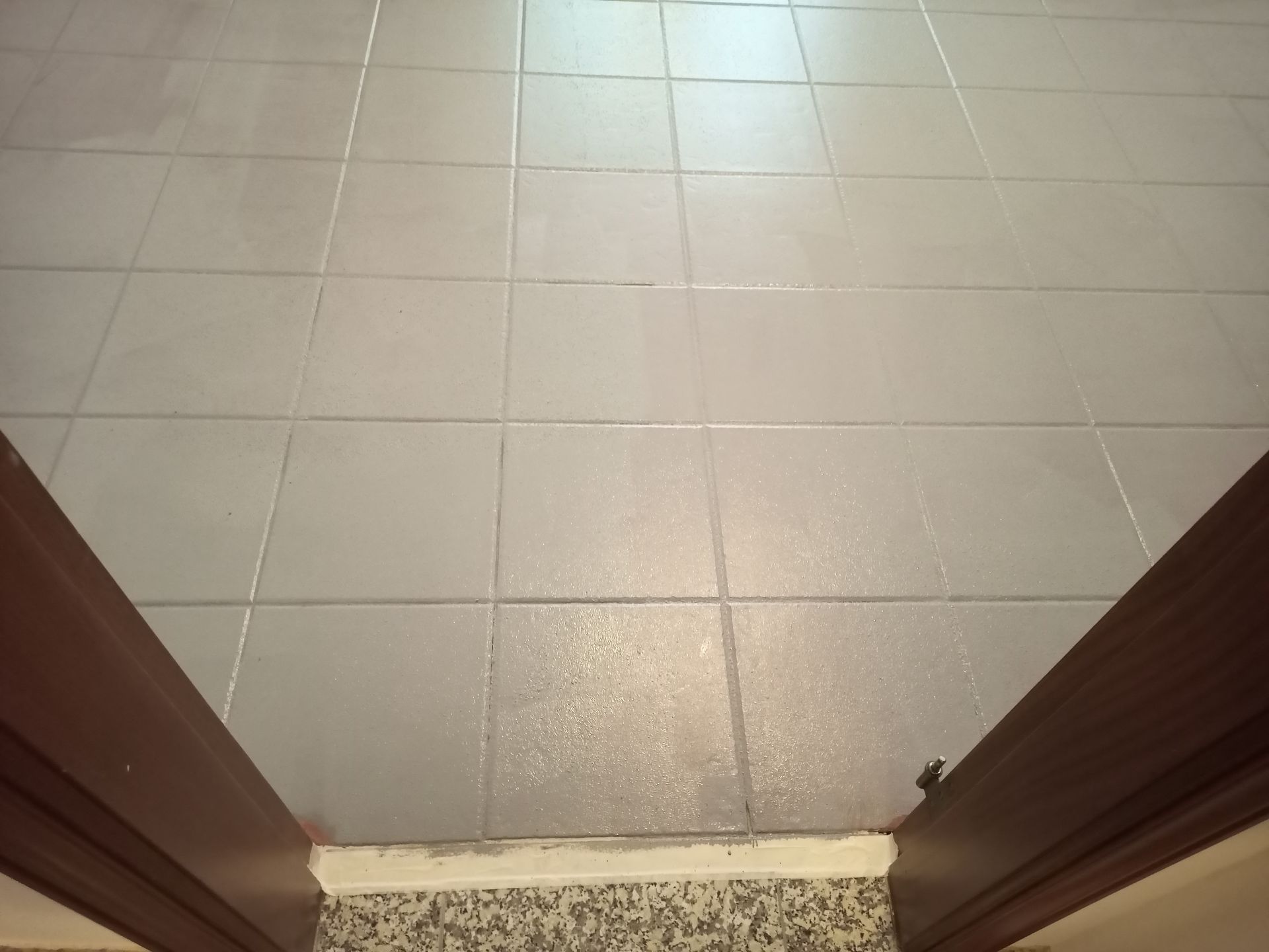 Piso de azulejos com detalhes em marrom escuro nas bordas inferiores. A luz reflete nos azulejos molhados.