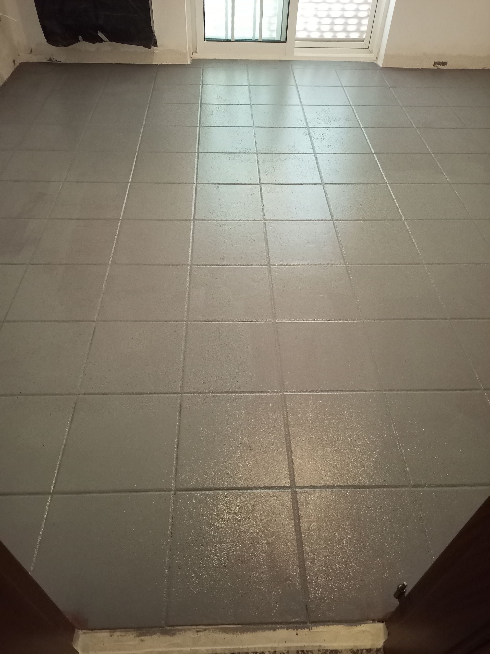 Piso de azulejos cinza em um cômodo, com uma moldura de porta na parte inferior e uma janela ao fundo.
