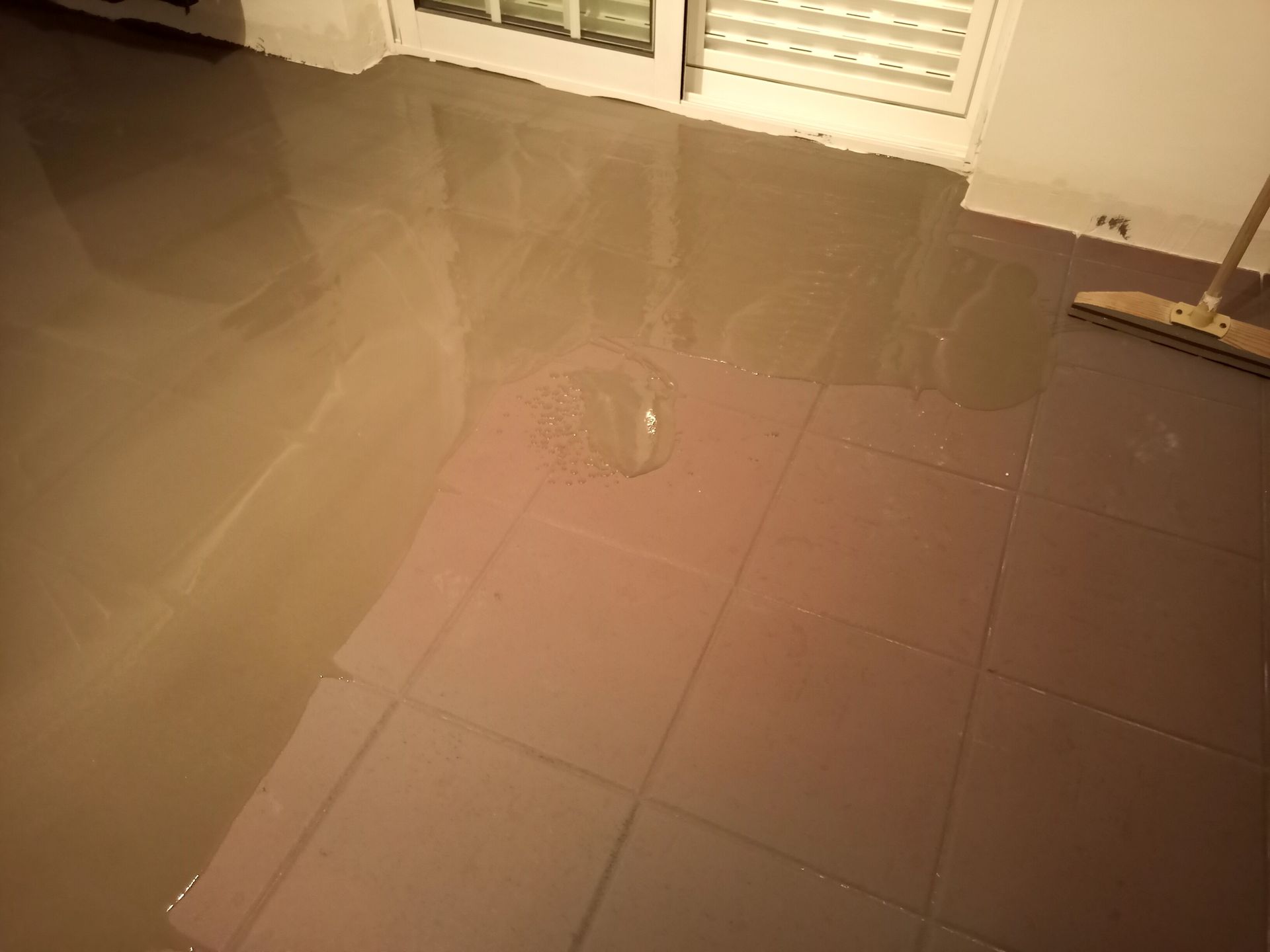Piso alagado com azulejos parcialmente submersos em um líquido cinza, provavelmente água. Um rodo está à direita.
