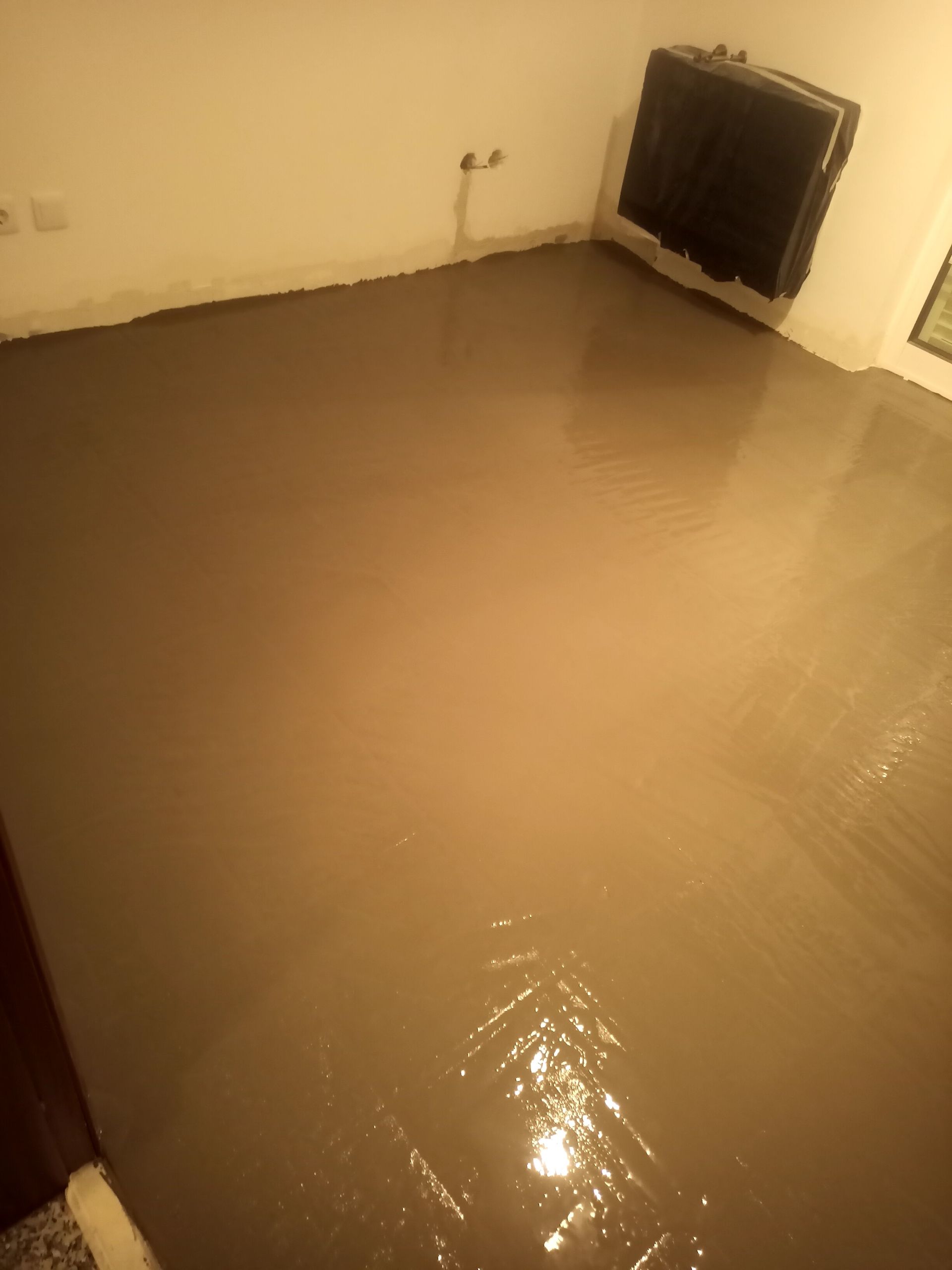 Piso autonivelante castanho-claro, recém-colocado e ainda úmido, em um cômodo, refletindo a luz. Paredes brancas, radiador preto.