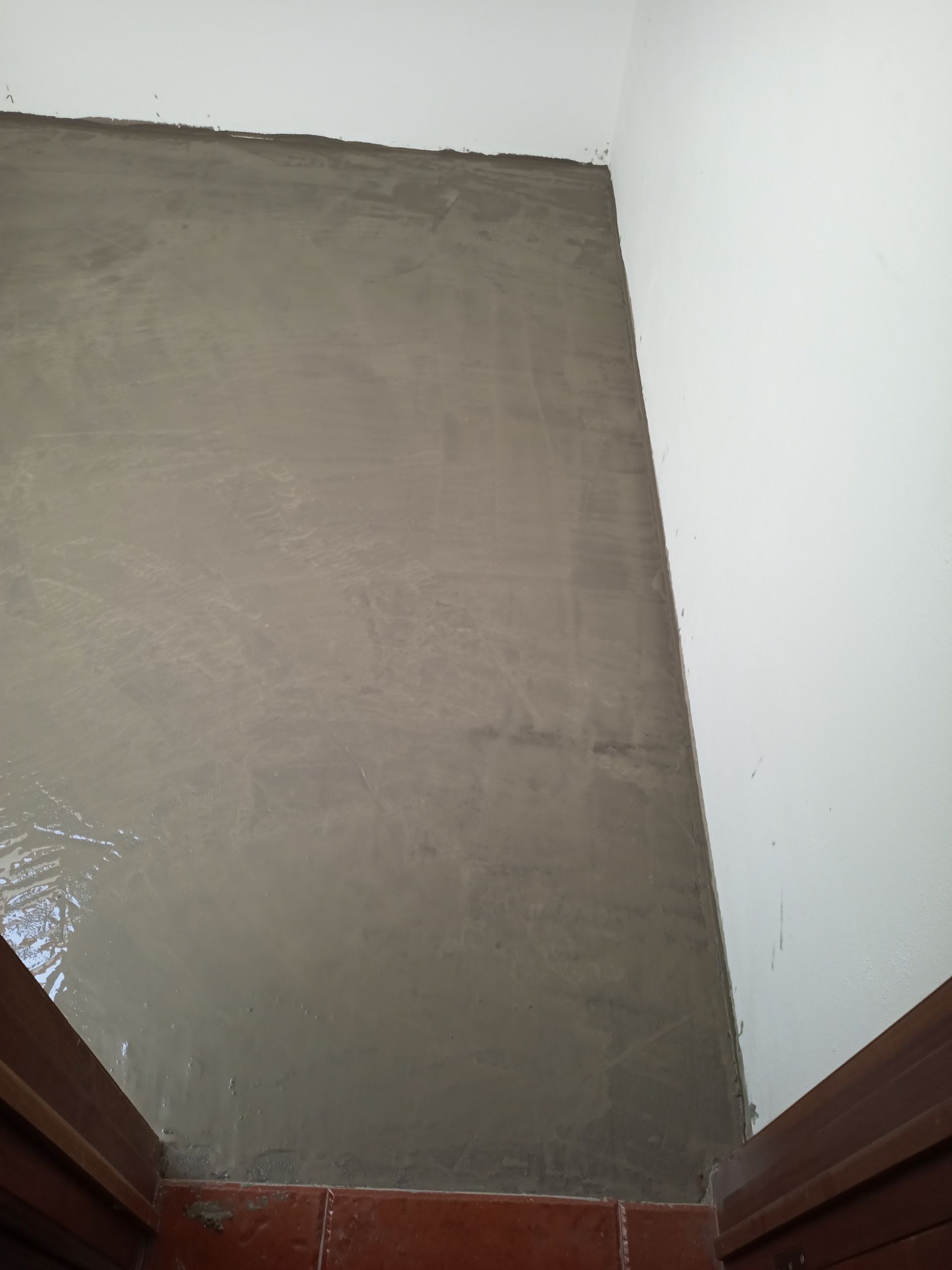 Piso de cimento cinza recém-assentado, contra paredes brancas e batente de porta marrom.