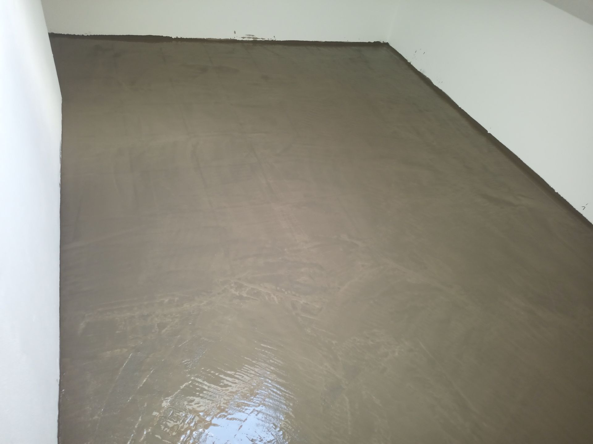 Piso de concreto recém-moldado em um cômodo, de cor cinza claro. Bordas próximas às paredes brancas.