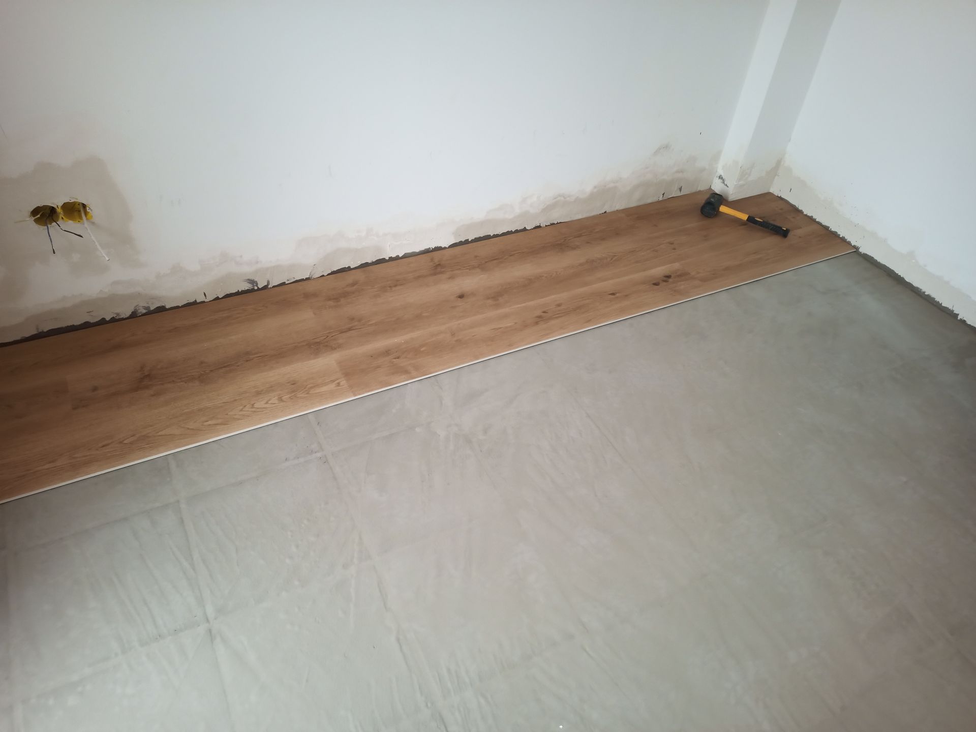 Instalação de piso vinílico imitando madeira em um cômodo. Uma fileira parcial de tábuas é instalada ao longo de uma parede, com as ferramentas à mostra.