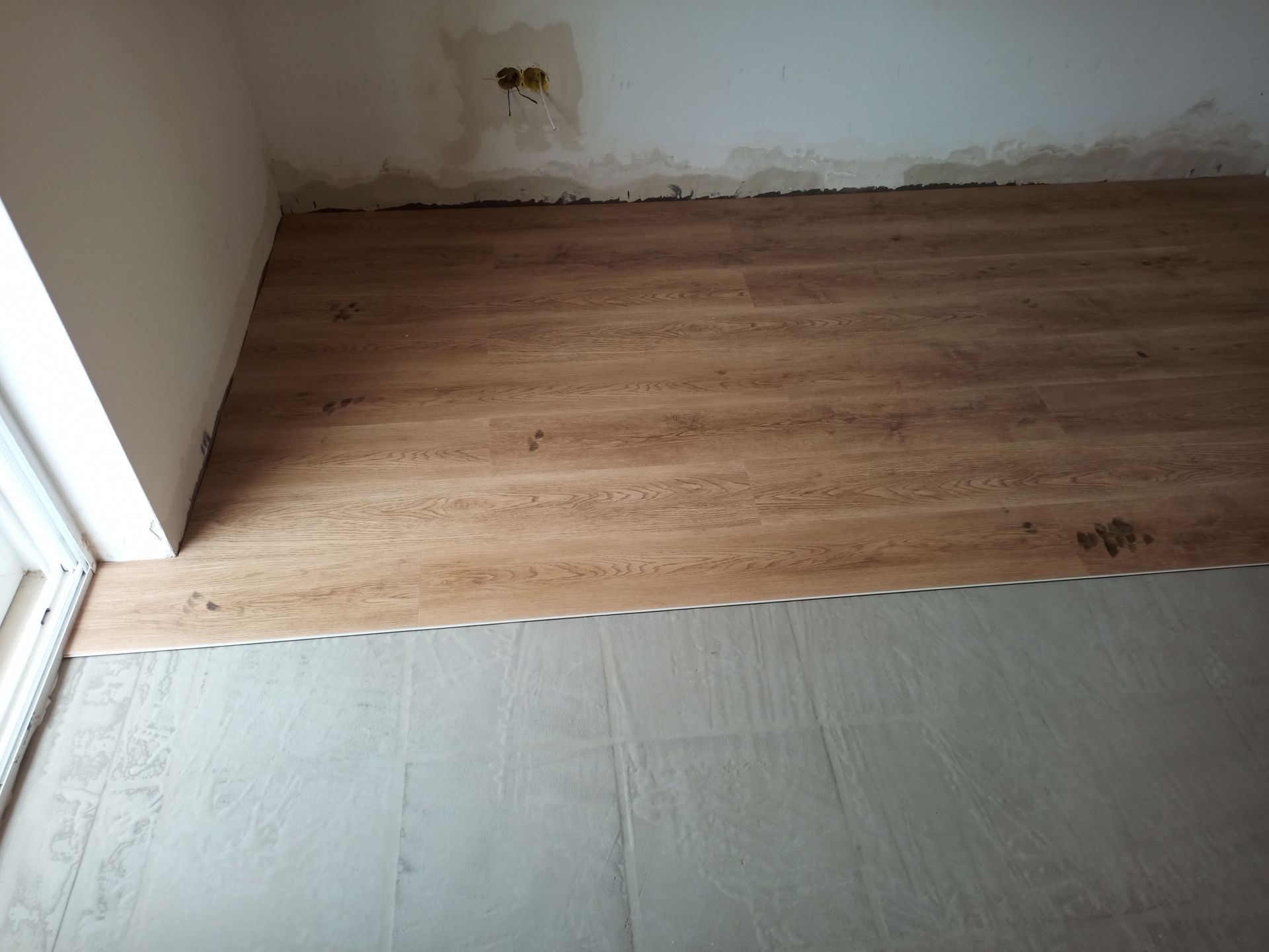 Piso com aparência de madeira sendo instalado sobre um contrapiso cinza em um cômodo com paredes brancas.