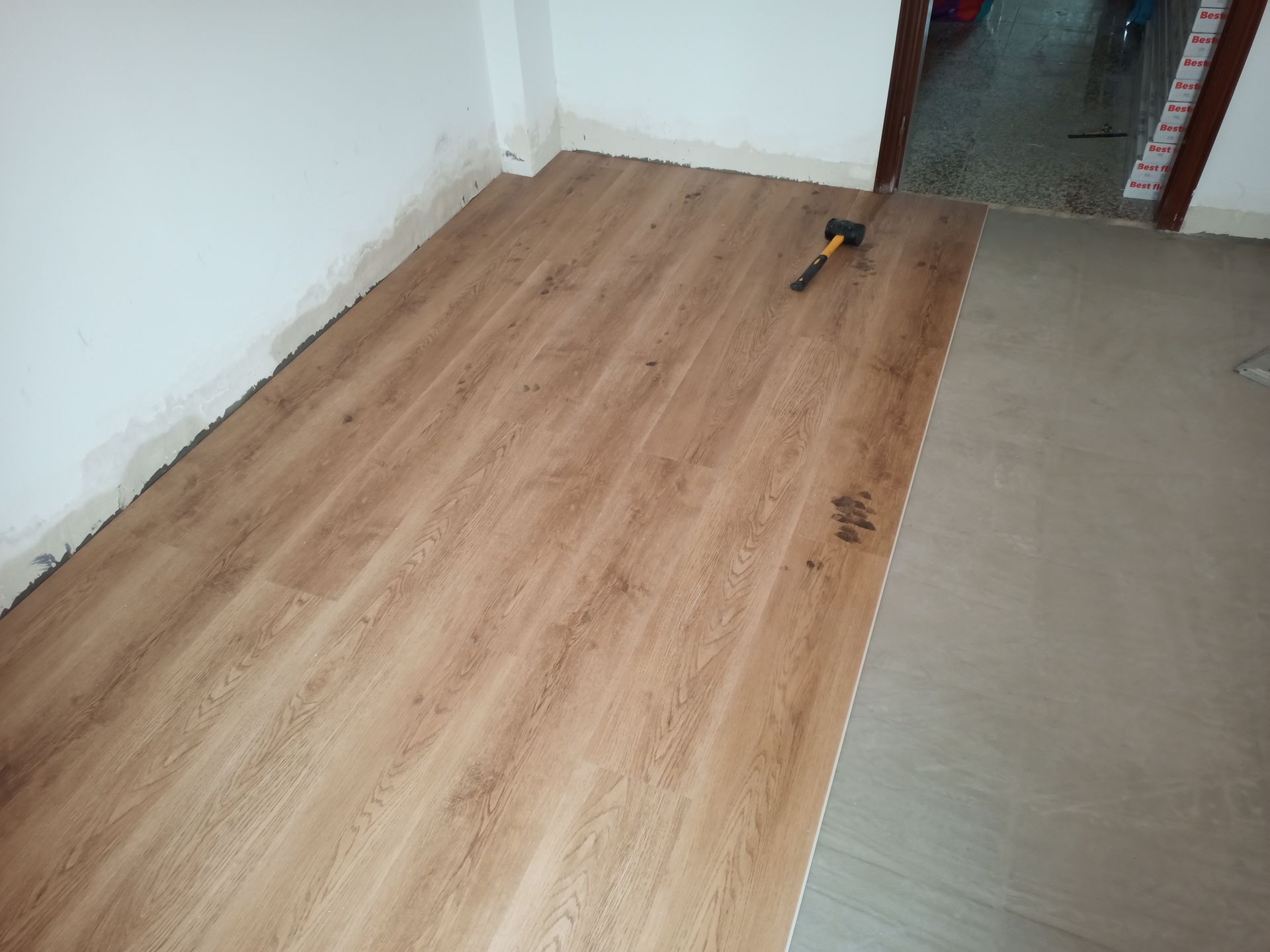 Instalação de piso vinílico imitando madeira em um cômodo. Um martelo está sobre as tábuas recém-instaladas. As paredes do cômodo são brancas.