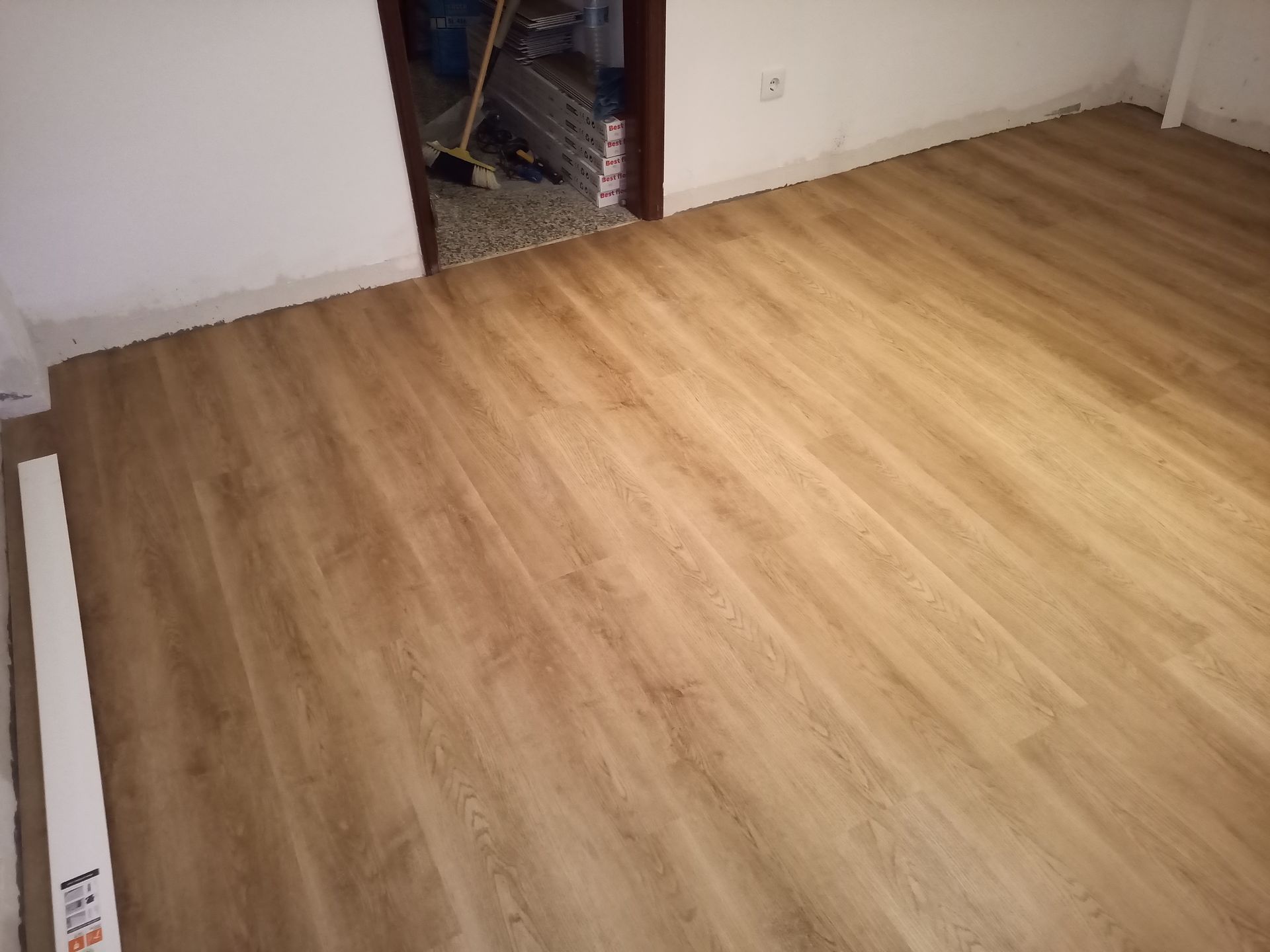 Piso imitando madeira em tom castanho claro instalado em um cômodo. Uma porta dá acesso a outro ambiente. Paredes brancas.