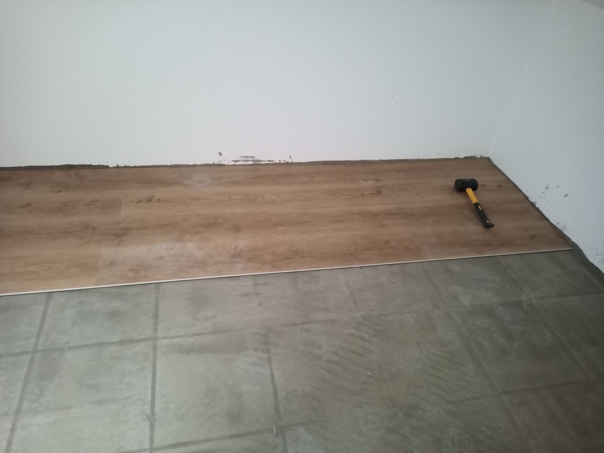 Instalação de piso imitando madeira sobre azulejos cinza existentes; um martelo está apoiado no novo piso próximo a uma parede branca.