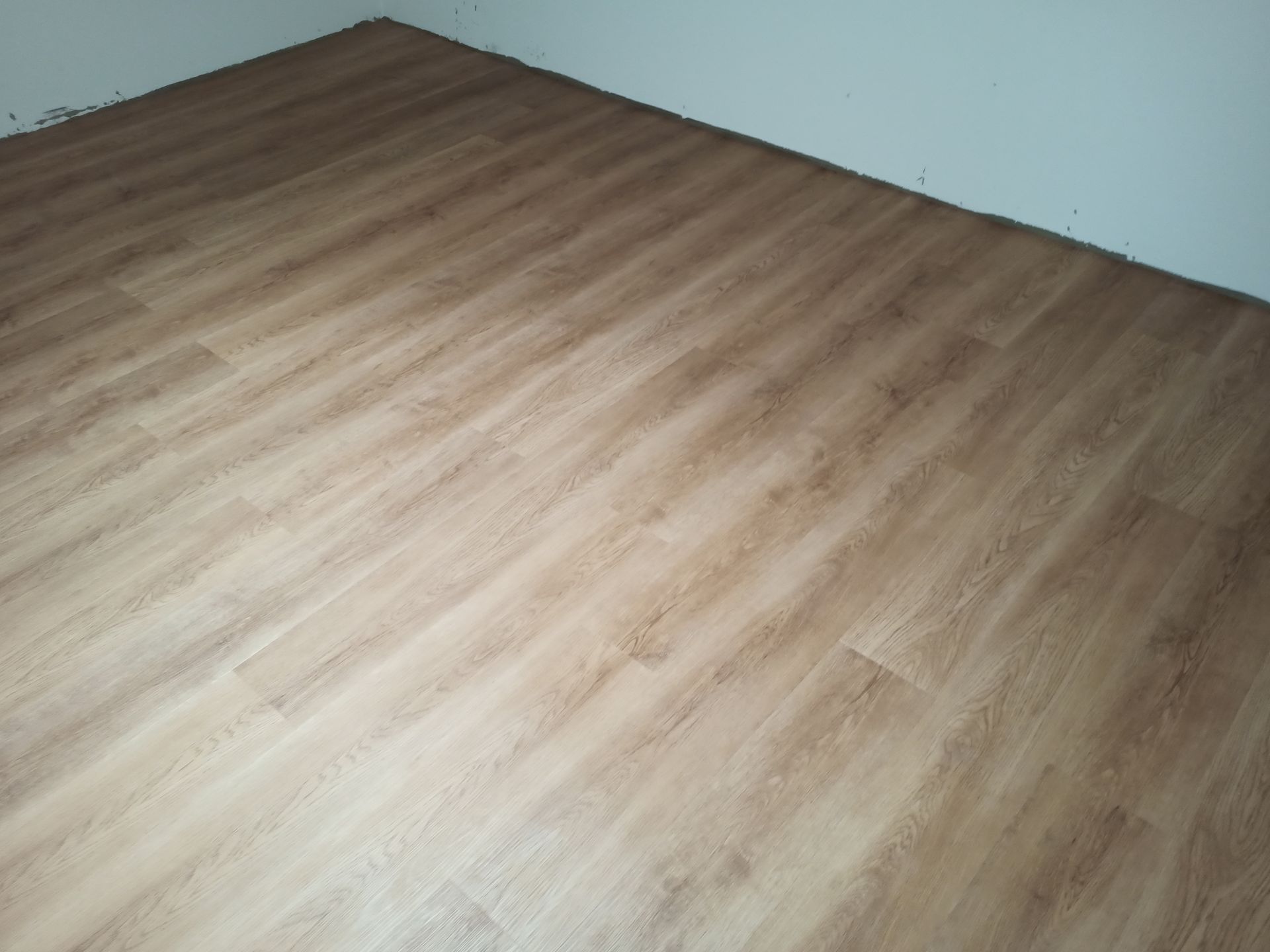 Piso imitando madeira em tom marrom claro instalado em um cômodo. As tábuas do piso são dispostas longitudinalmente.