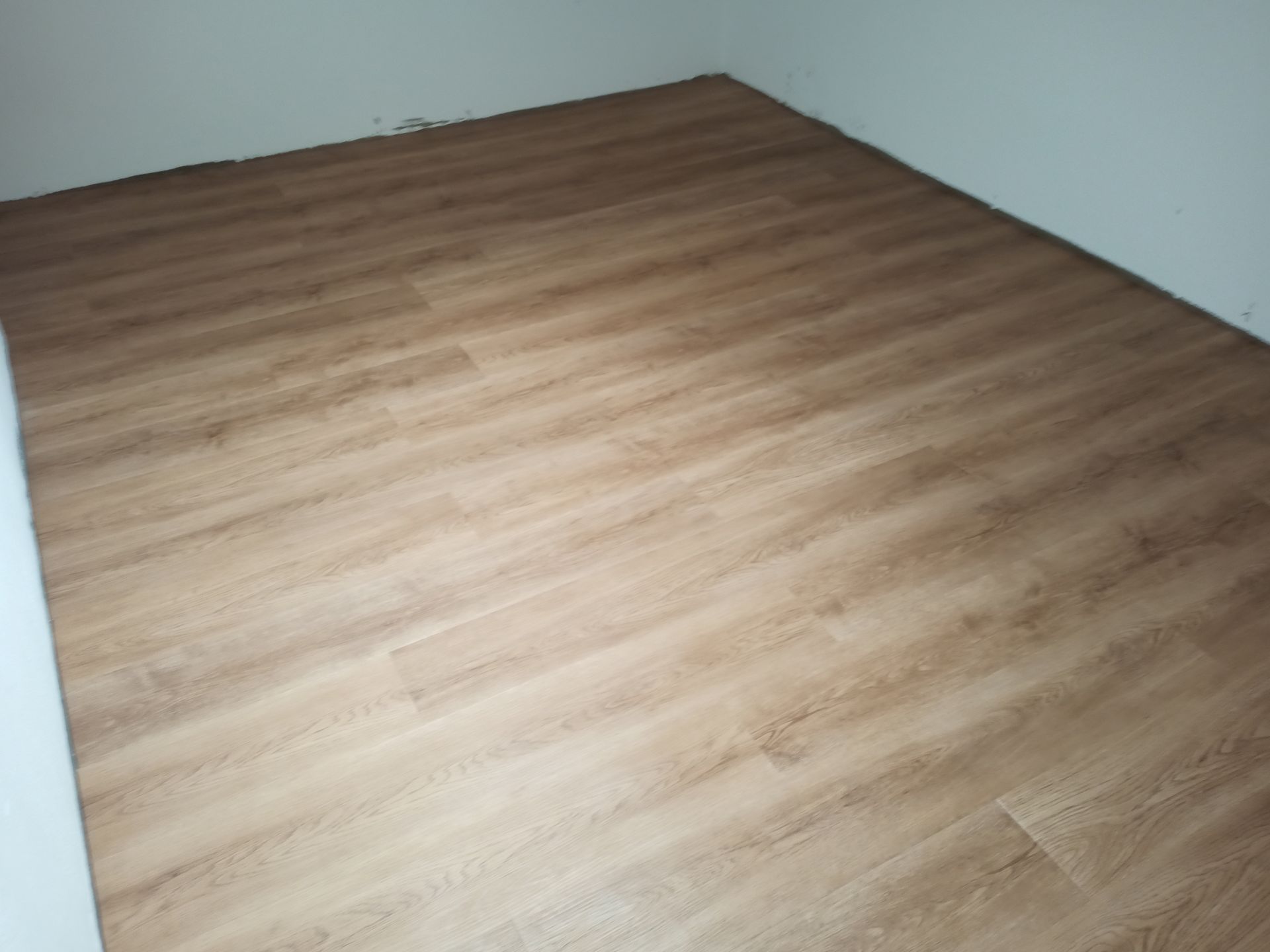 Quarto vazio com piso que imita madeira em tom castanho claro. Paredes brancas são visíveis no canto.