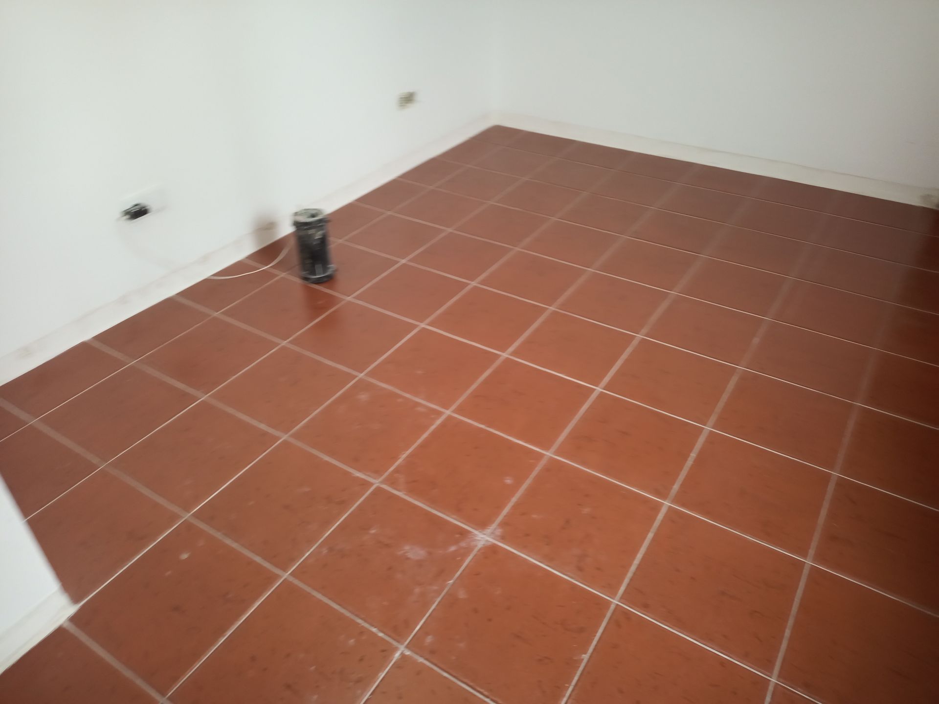 Piso de azulejos marrons em um cômodo com paredes brancas. Um pequeno recipiente escuro está sobre o chão.