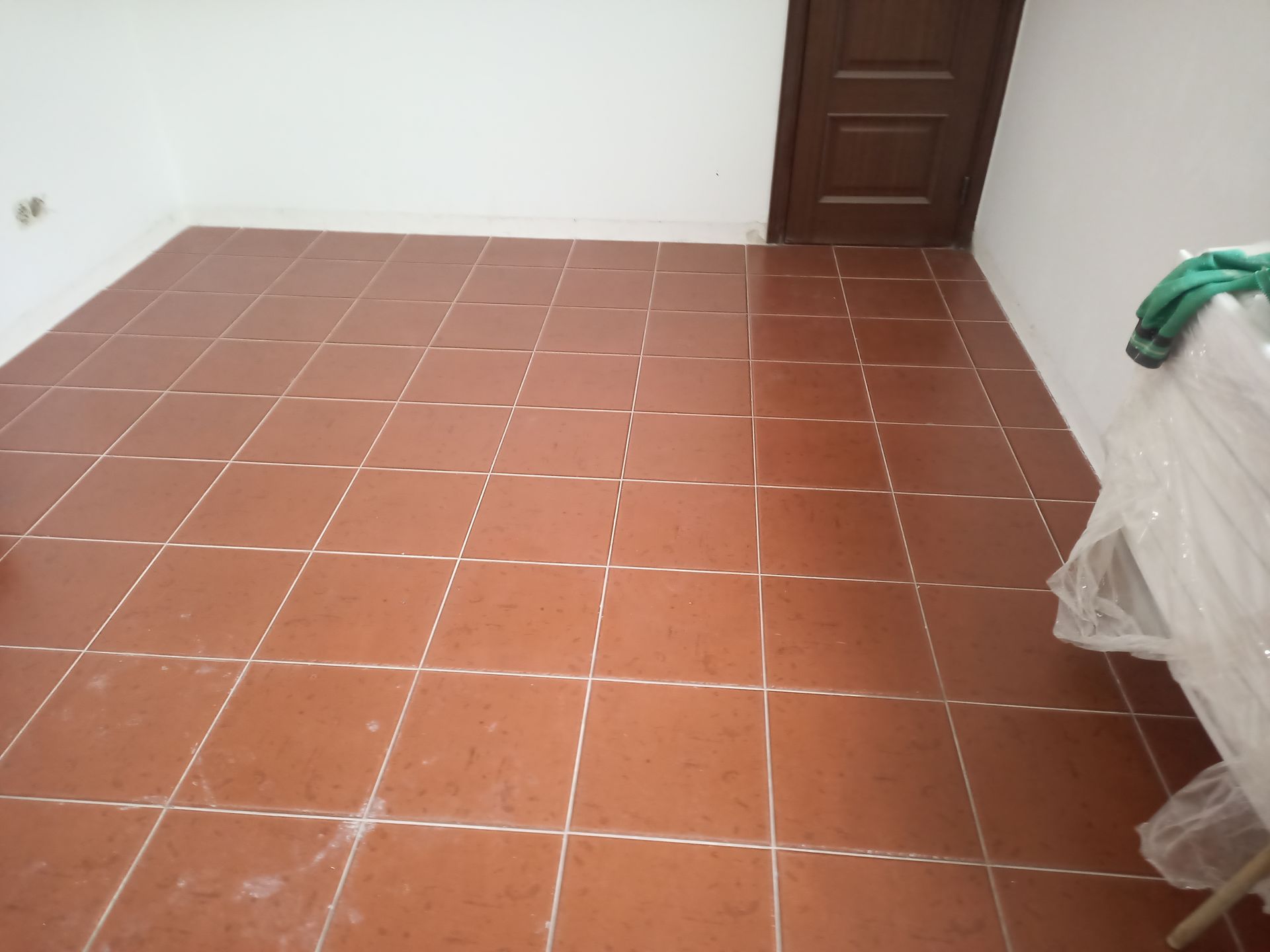 Piso de azulejo marrom em um quarto com paredes brancas e porta marrom; algumas roupas estão em um canto.