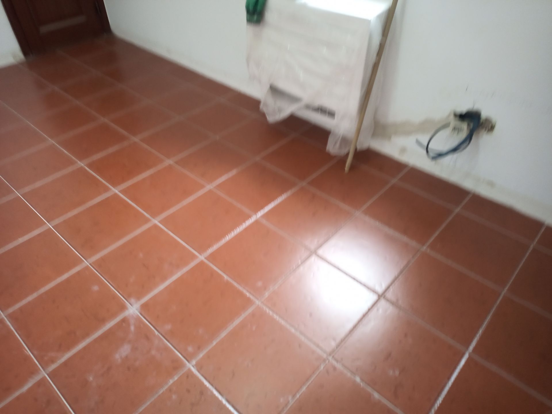 Piso de azulejo marrom em um cômodo com paredes brancas, um objeto coberto e uma tomada elétrica.
