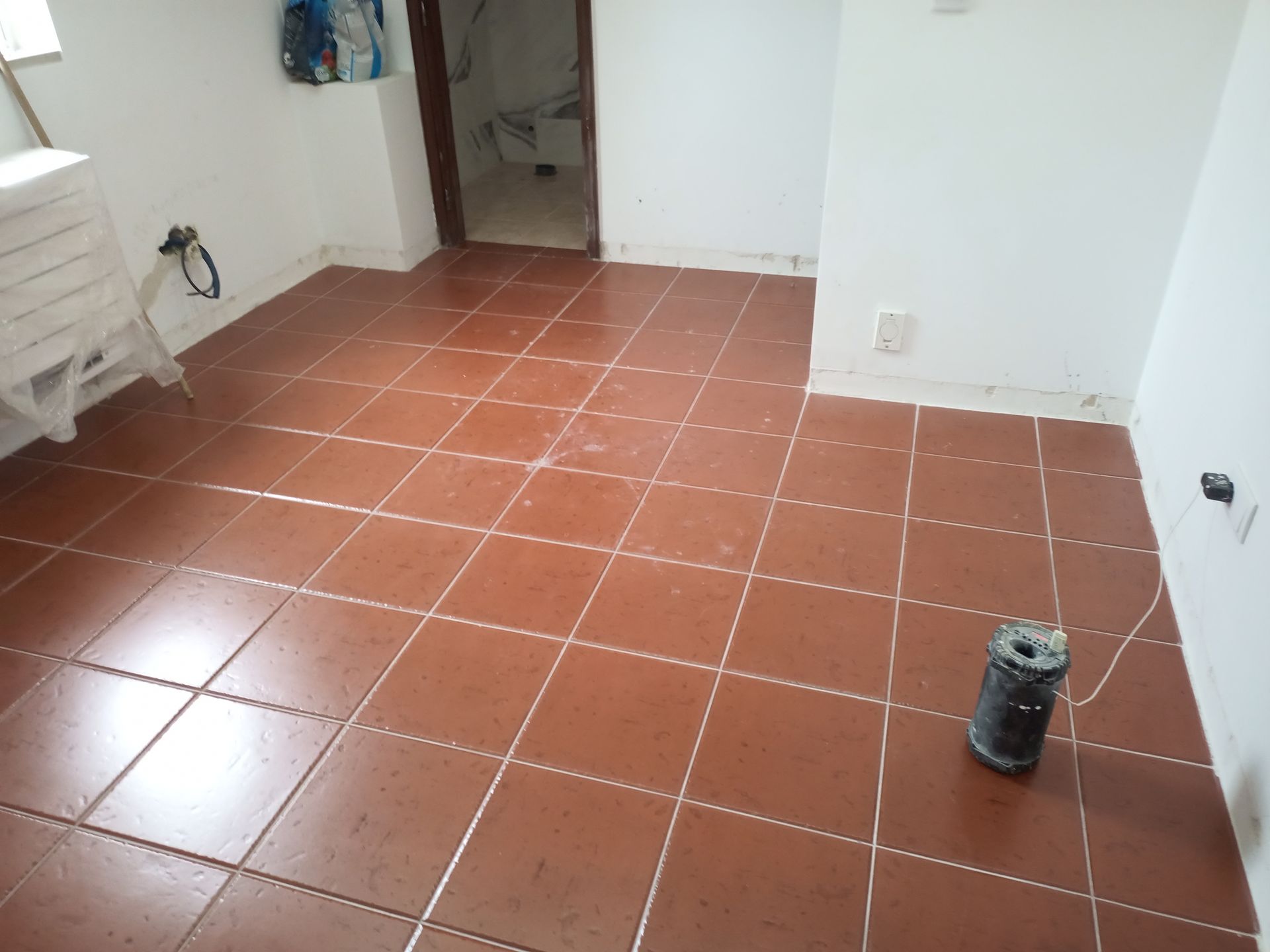 Piso de ladrilho vermelho em uma sala com paredes brancas, uma porta e um recipiente preto.
