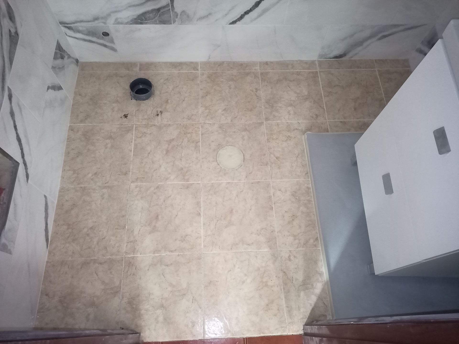 Piso do banheiro revestido com azulejos bege, com ralo para vaso sanitário e armário branco.