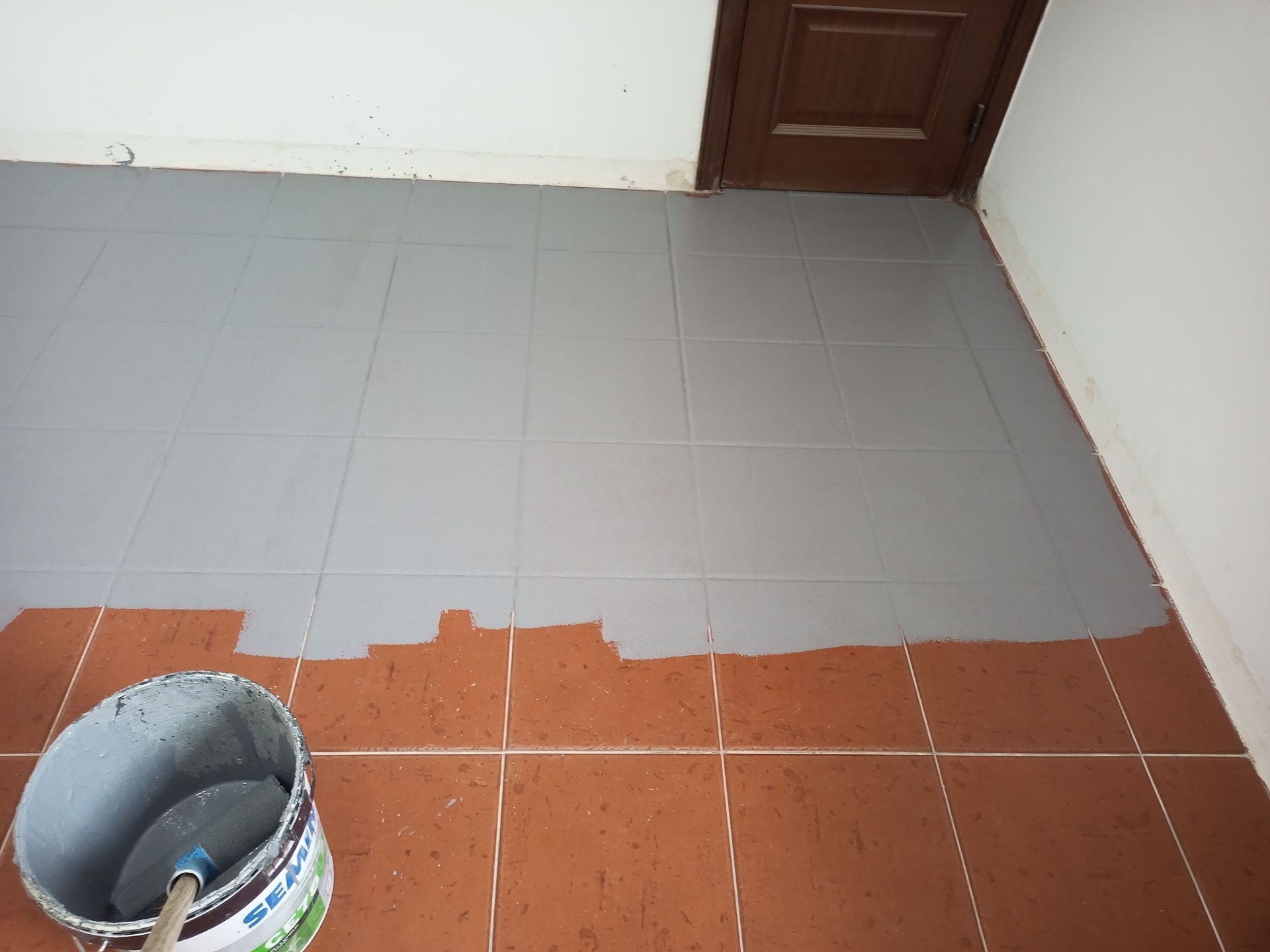 Tinta cinza sendo aplicada nos ladrilhos do chão, com balde e espátula à vista. Ladrilhos marrons parcialmente pintados.
