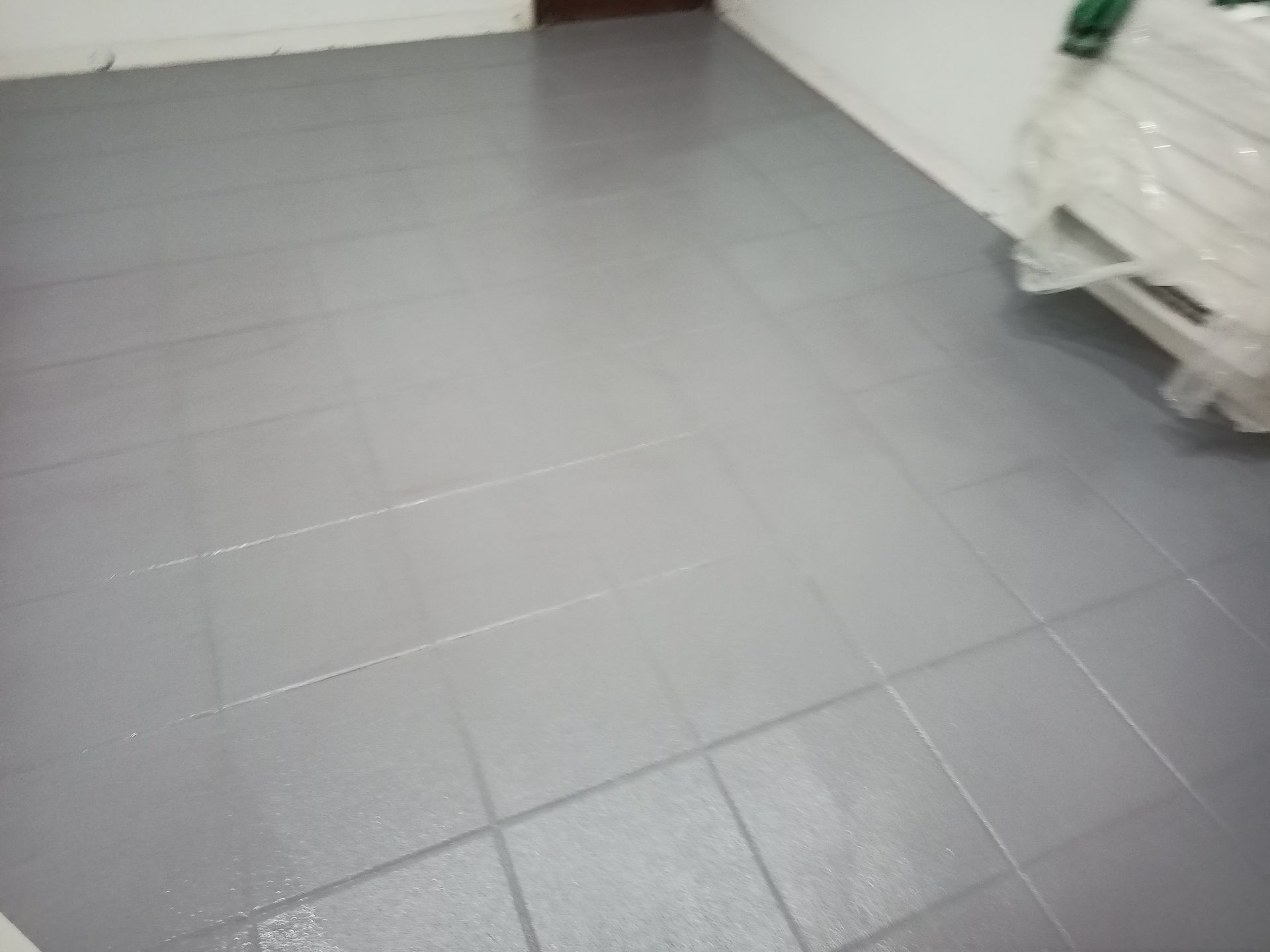 Piso de azulejos cinza, parcialmente molhado, refletindo a luz. Canto do cômodo com paredes brancas e uma porta.