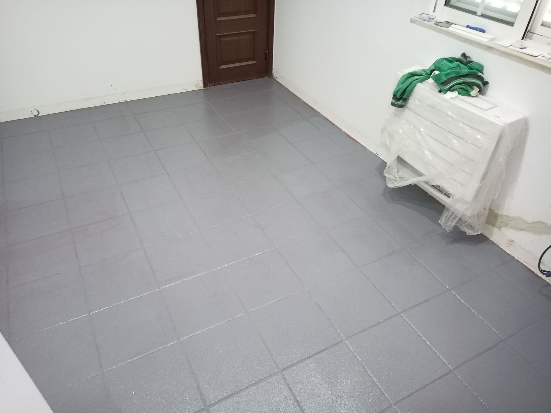 Piso de azulejo cinza em um cômodo com paredes brancas, porta marrom e um eletrodoméstico embalado.