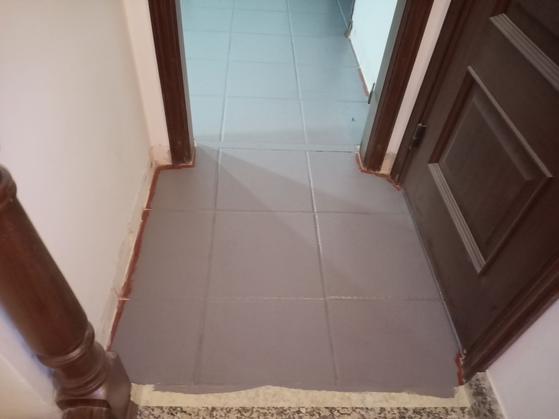 Corredor com piso de ladrilho cinza, portas e molduras marrons e corrimão de madeira.