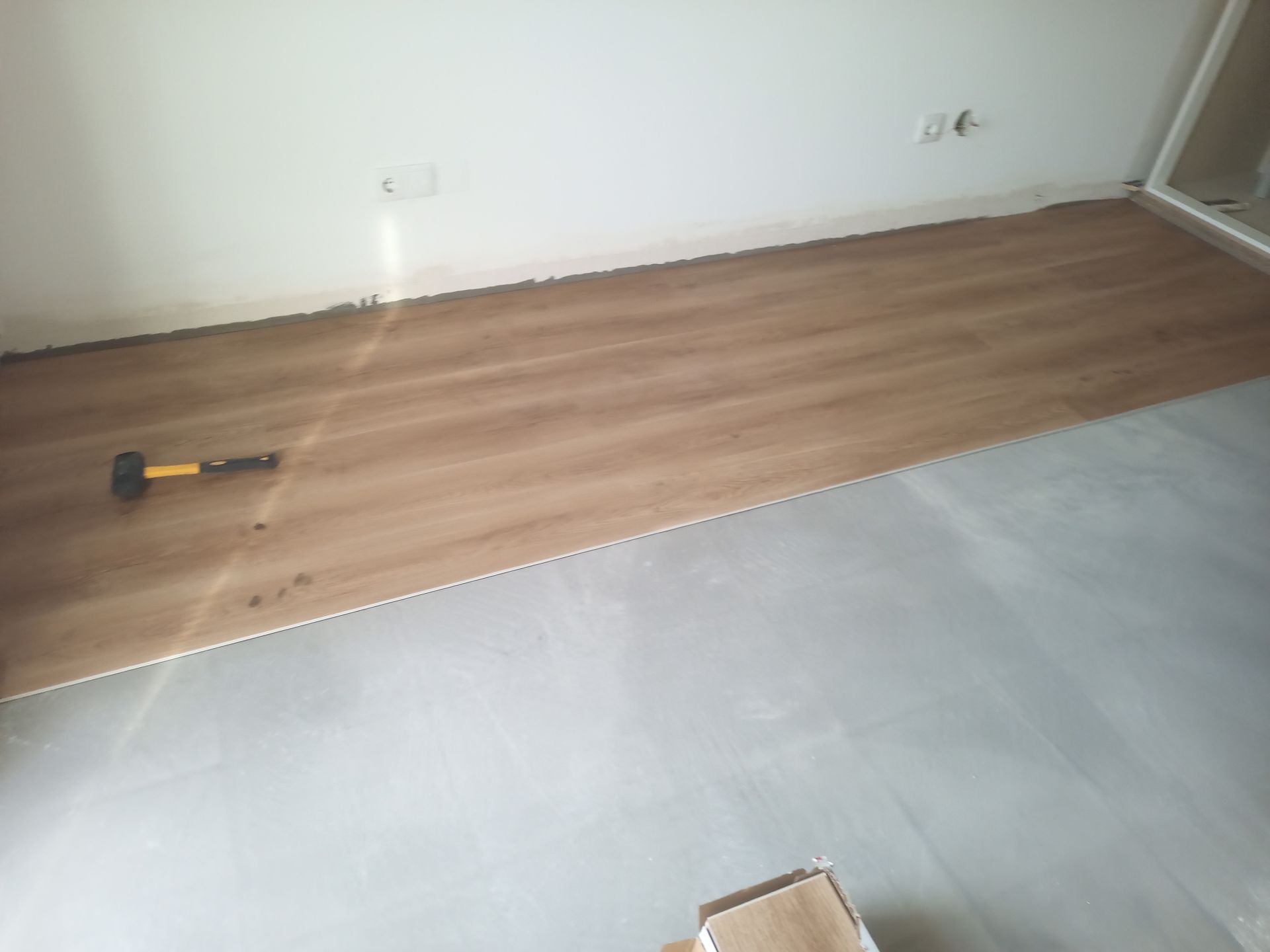 Piso de madeira instalado sobre uma base cinza em um cômodo com paredes brancas. Um martelo está sobre as tábuas.