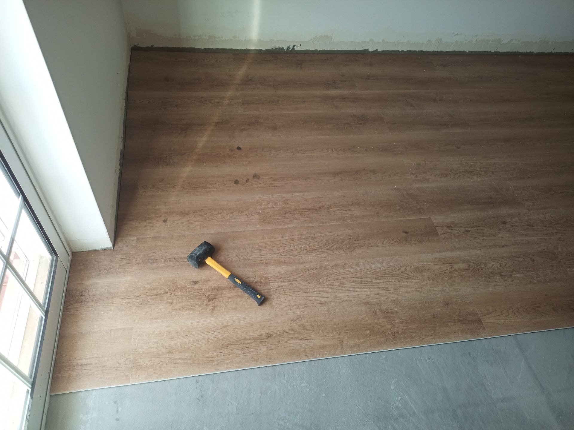 Instalação de piso: aplicação de martelo em piso laminado com aparência de madeira parcialmente instalado em um cômodo com parede branca e janela.
