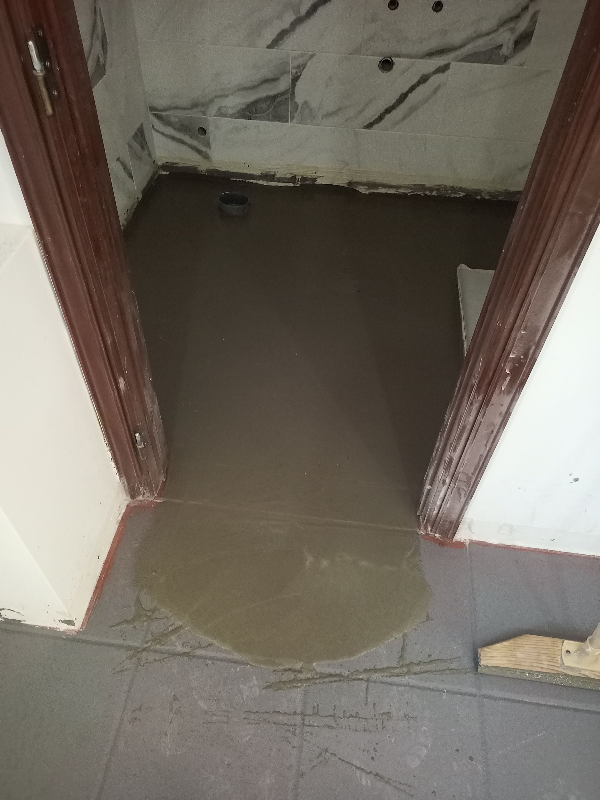 Piso do banheiro com concreto molhado na entrada. Batentes da porta em marrom escuro e parede branca.