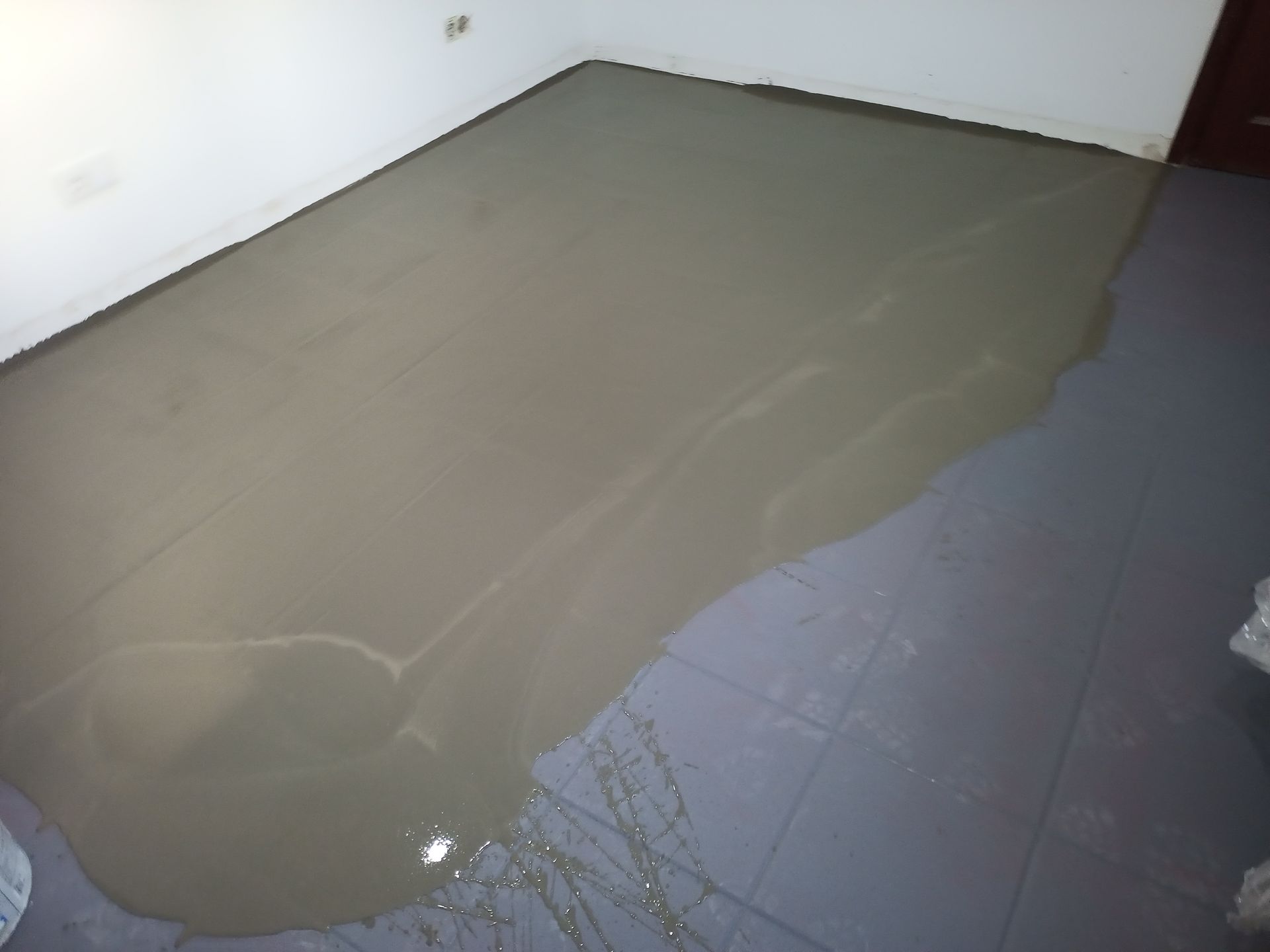 Cimento úmido derramado sobre o piso de azulejos escuros, transbordando da borda perto de uma parede branca.