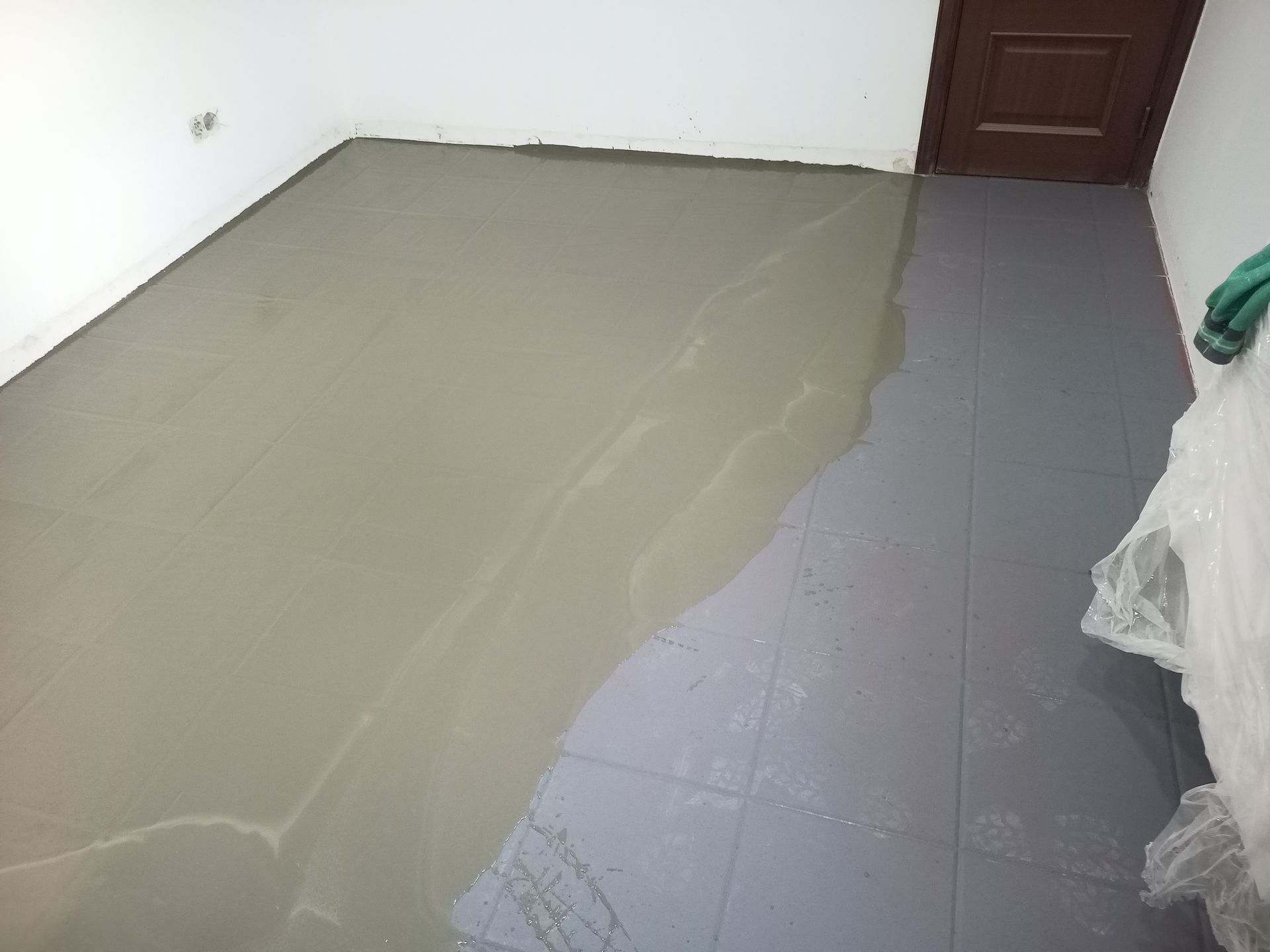 Piso cinza parcialmente coberto por um material cinza claro e úmido. Uma porta e paredes brancas são visíveis.