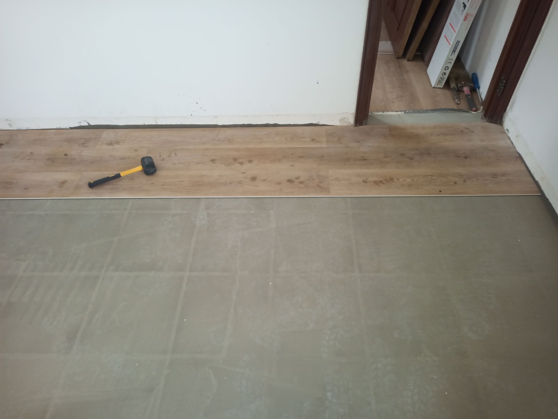 Transição de piso: tábuas de madeira clara encontrando azulejos cinza, separados por uma faixa metálica. Martelo apoiado nas tábuas.