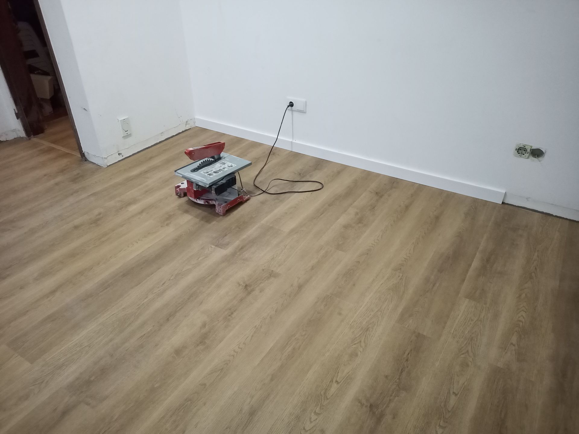 Uma pequena serra de mesa está sobre um piso de madeira novo em uma sala com paredes e rodapés brancos.