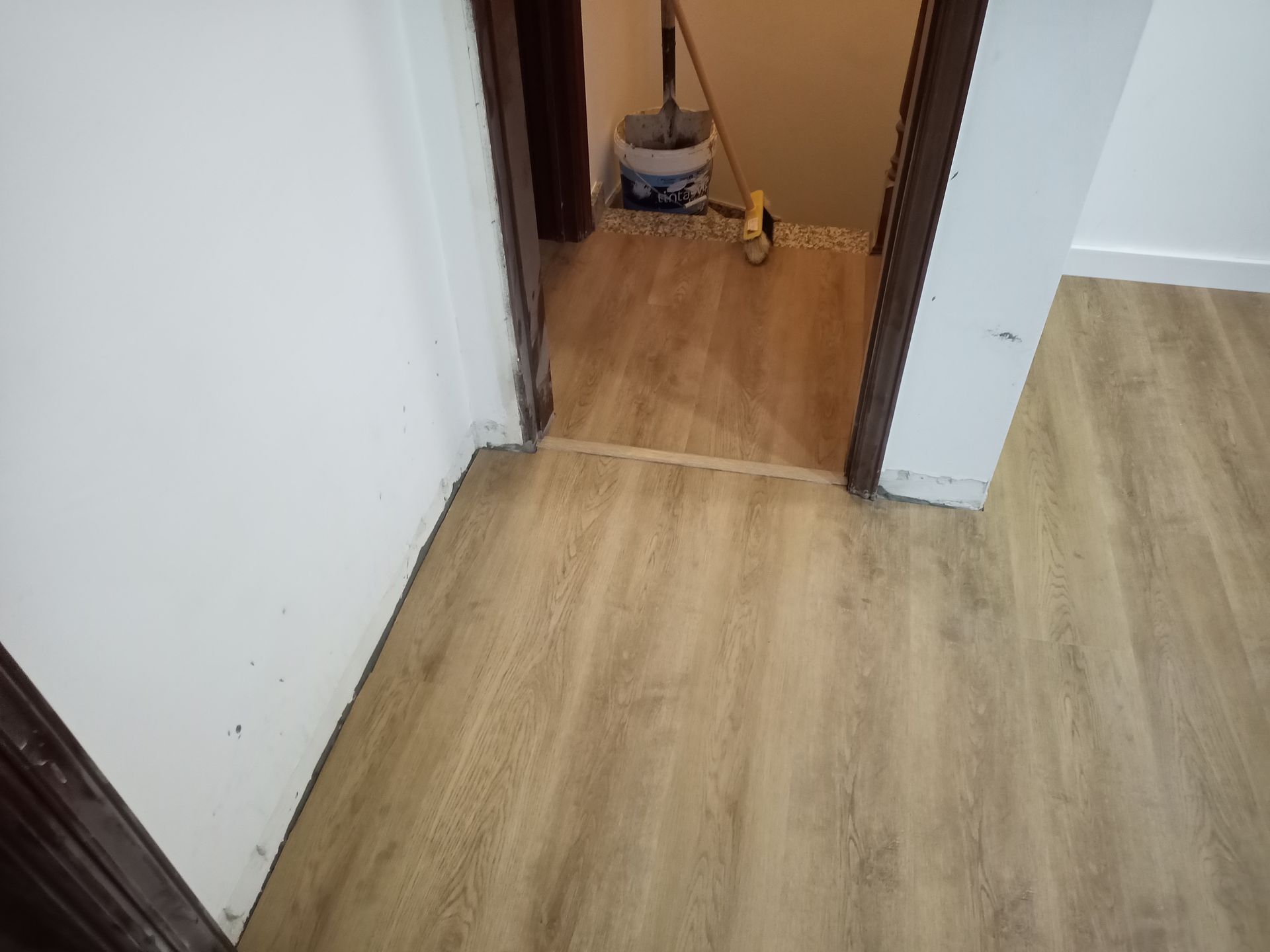 Vista de uma porta com piso que imita madeira clara e um batente mais escuro. Um esfregão e um balde são visíveis dentro da porta.