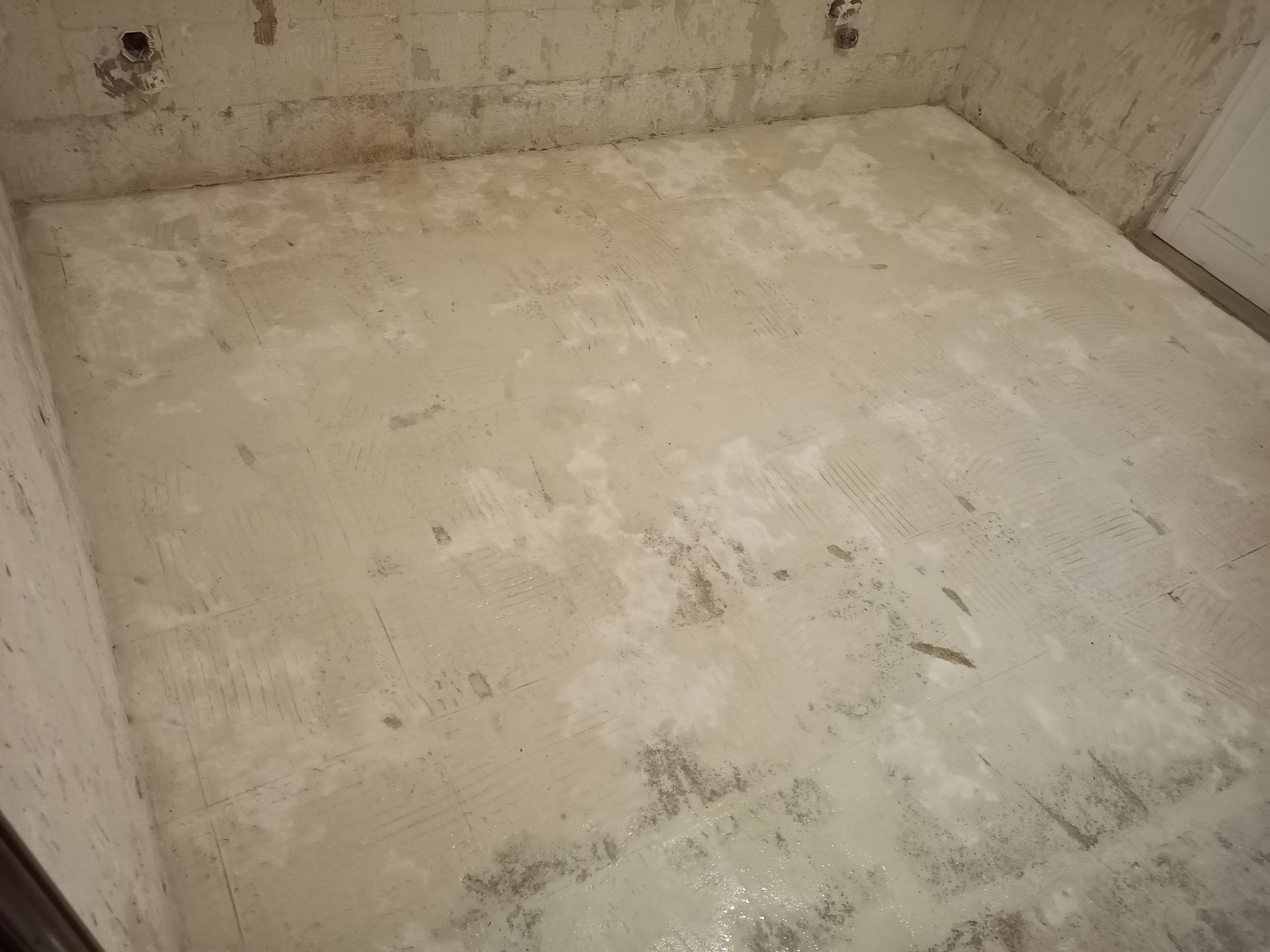 Sala de concreto com piso irregular de cor clara e paredes de concreto. Uma porta branca é visível.