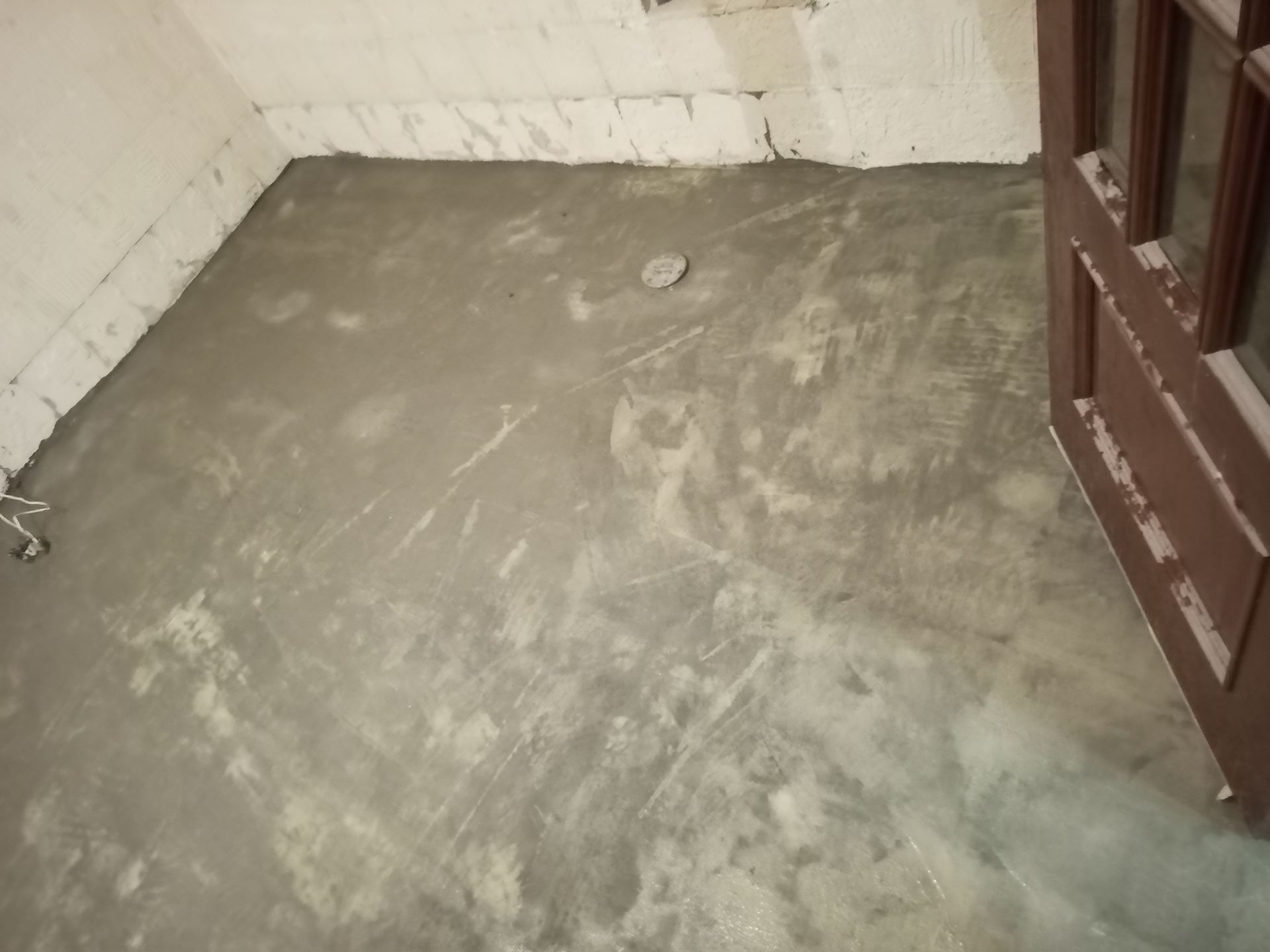 Piso de cimento recém-colocado em um cômodo. Batente de porta marrom à direita. Paredes cinza e branca.