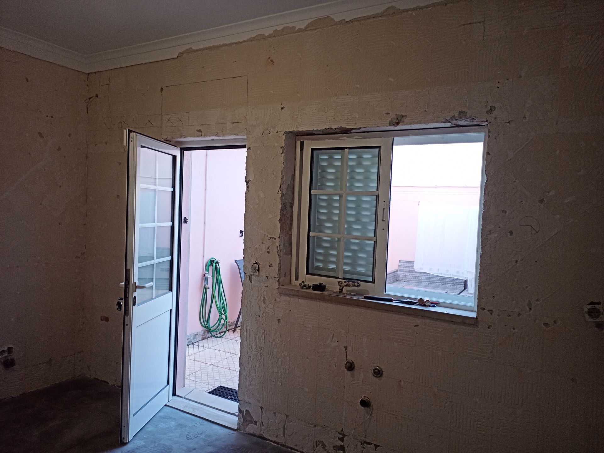Quarto interior com porta e janela abertas; paredes inacabadas, caixilhos de porta e janela brancos.