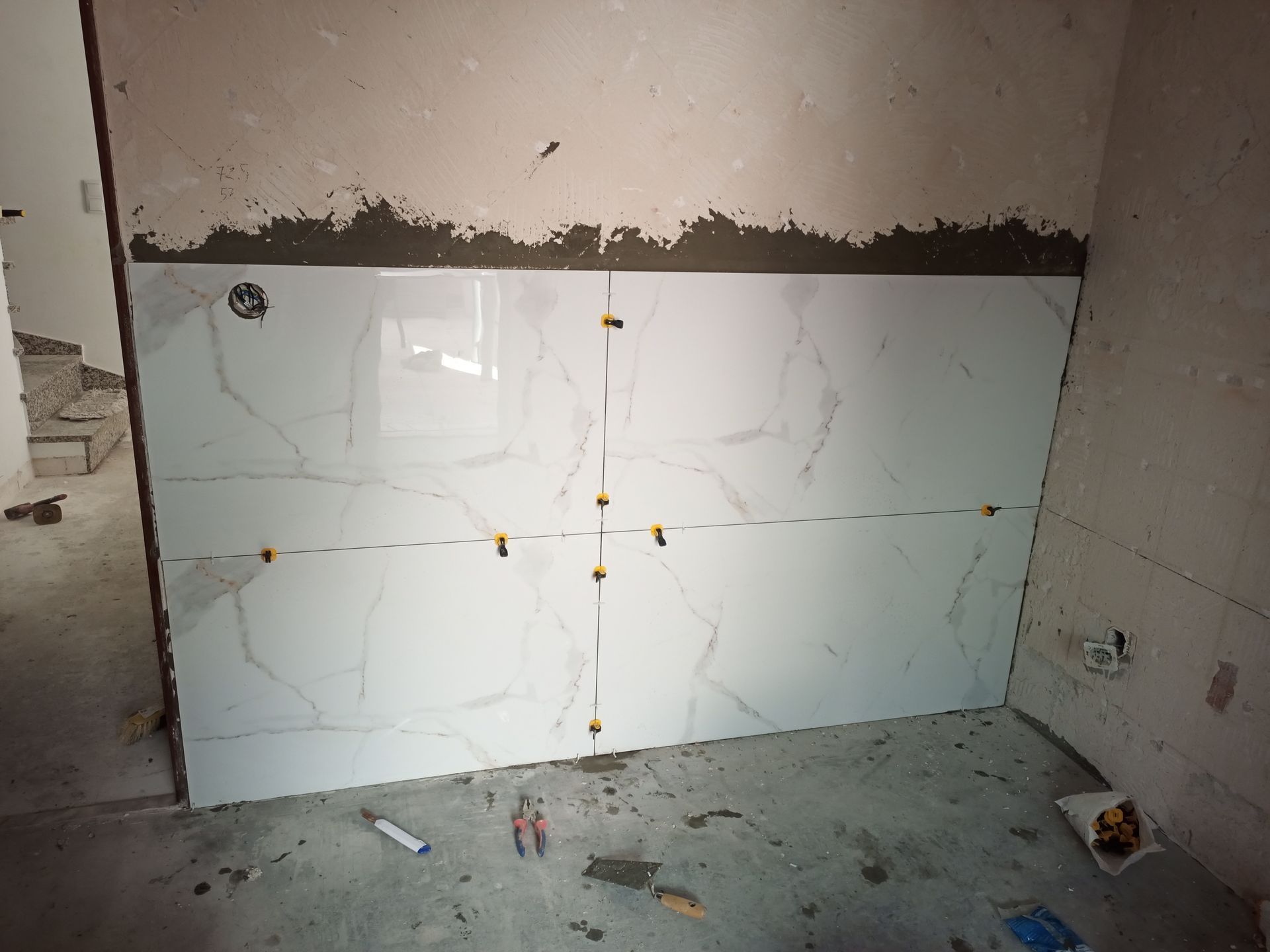 Azulejos brancos parcialmente instalados em uma parede. Espaçadores amarelos são visíveis. Ambiente de construção.