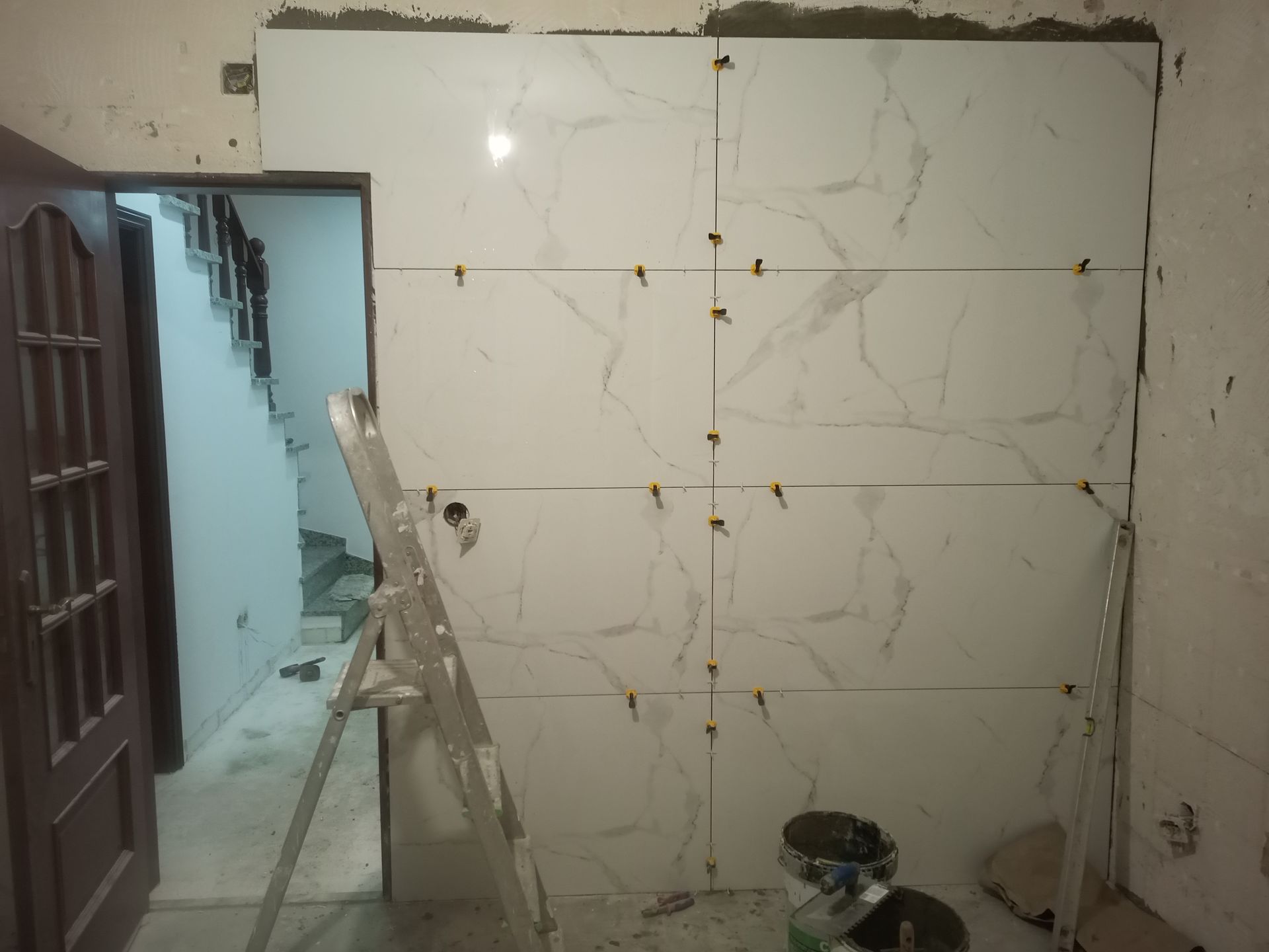 Parede com grandes azulejos brancos sendo instalados, escada, porta e degraus visíveis.