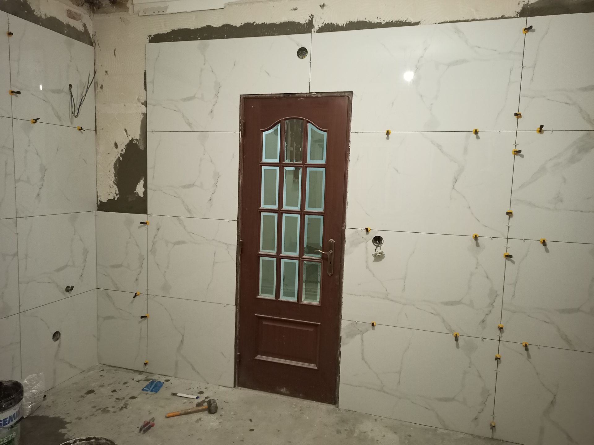 Sala com instalação de azulejos brancos imitando mármore. Uma porta marrom está centralizada. Espaçadores e entulho de construção presentes.