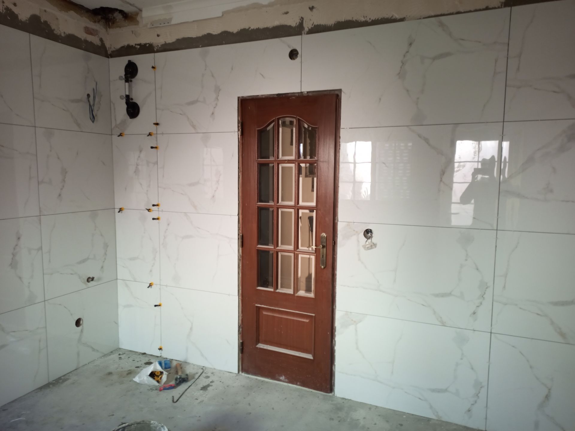 Quarto com paredes revestidas de azulejos brancos, porta marrom e teto inacabado.